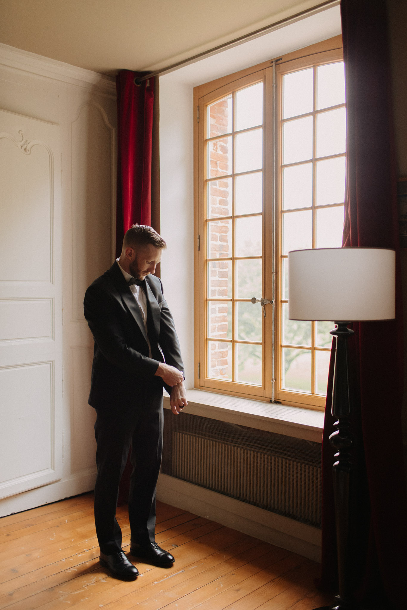 The groom is getting ready in an indoor room, adjusting his cufflinks or shirt sleeve while looking down. He is wearing a dark navy suit with a white dress shirt and a dark tie, paired with black oxford shoes. The room has classic French architectural details including a white panelled door with ornate moulding, warm honey-toned hardwood floors, deep crimson velvet curtains, and a multi-pane window with a natural wood frame through which brick exterior walls are visible. A dark wood floor lamp with a cream drum shade stands to the right. The shot is a full-length portrait with natural window light illuminating the groom from the side.