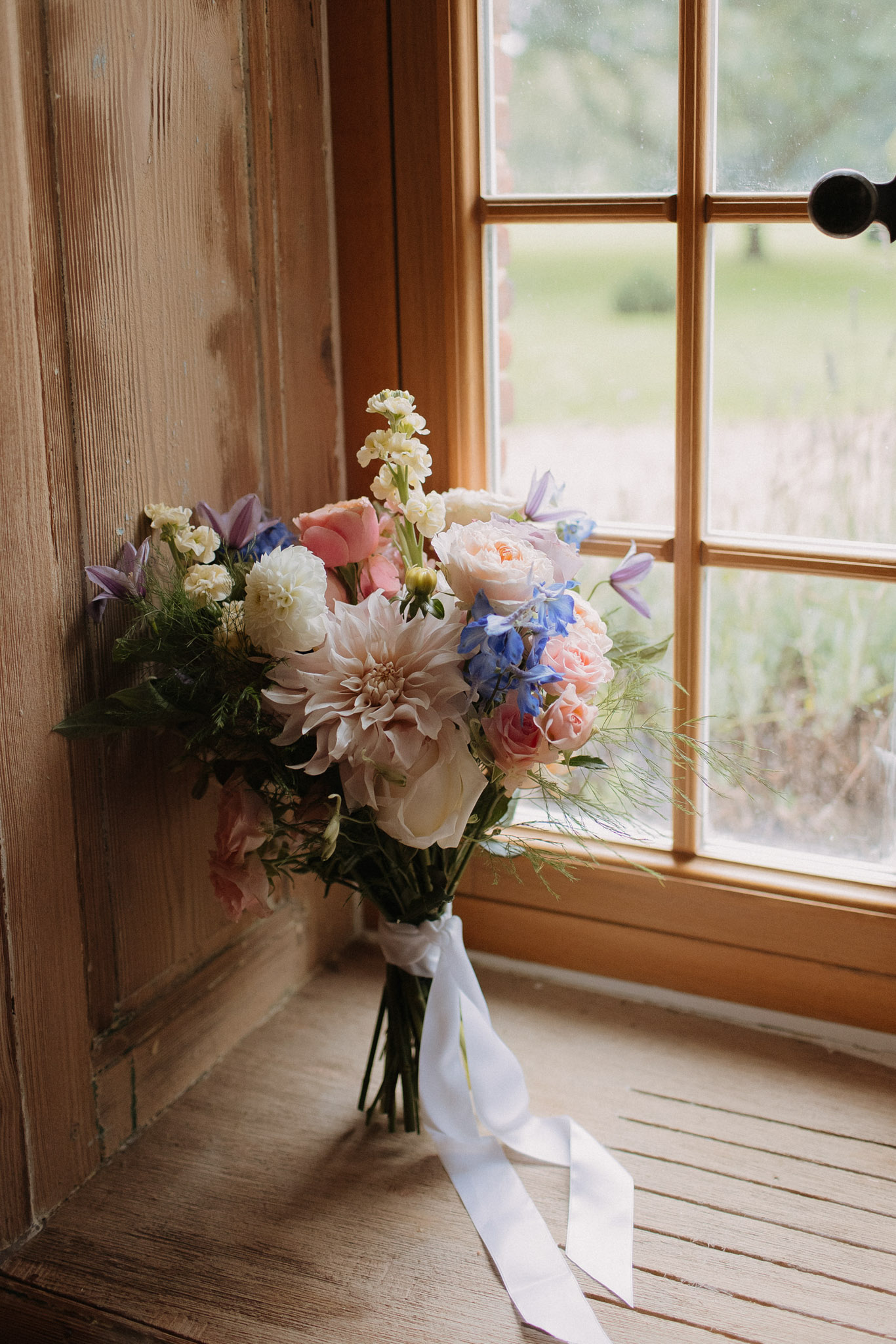 Garden-style bouquet with blush dahlia, peach roses, and blue delphinium tied with satin ribbon at window