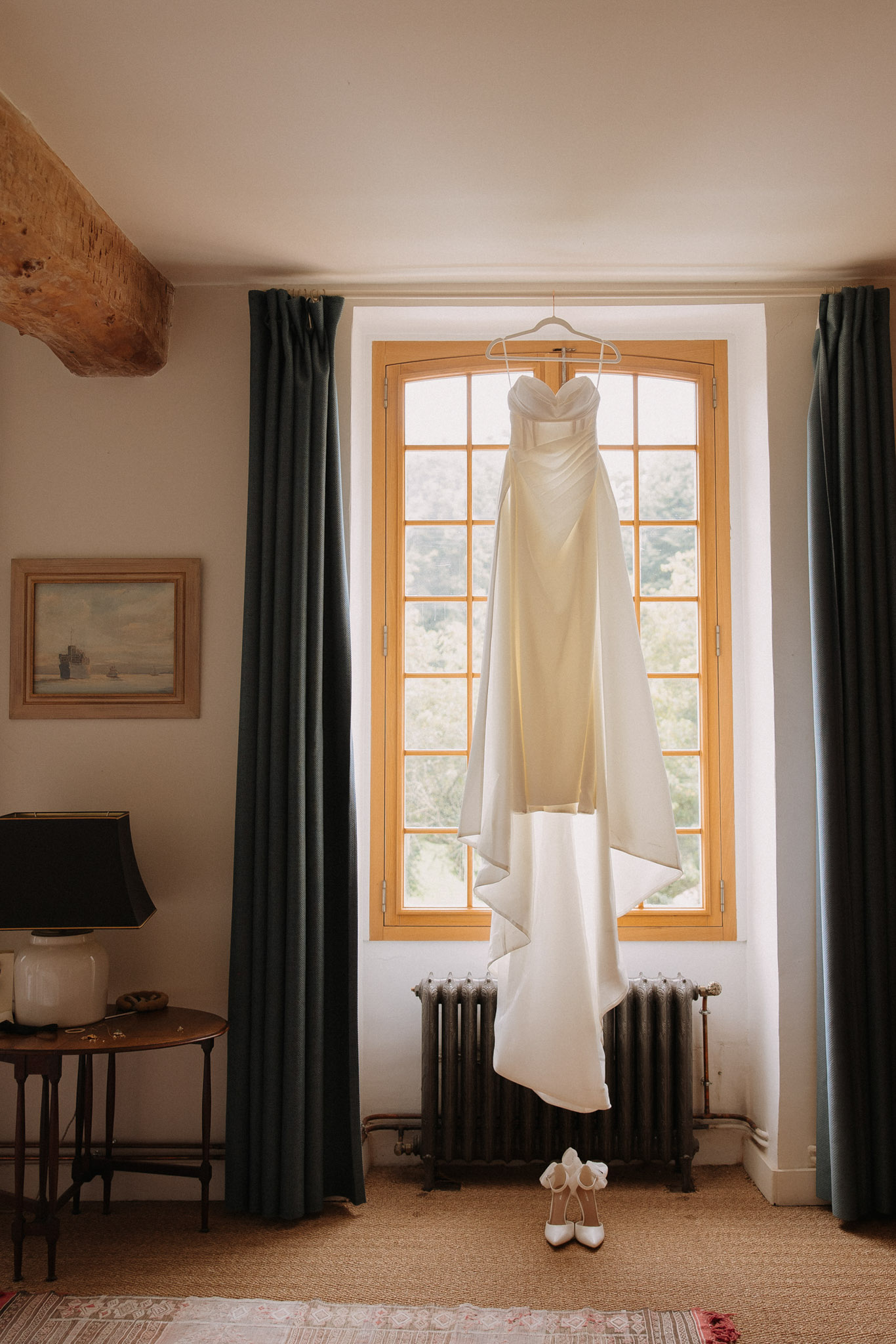 A getting-ready detail shot taken indoors in what appears to be a château bedroom or suite. An ivory strapless wedding gown with a sweetheart neckline, fitted silhouette, and a dramatic draped train is hung from a hanger in front of a tall arched window with warm honey-toned wooden frames. A pair of white satin heeled shoes with bow detailing is placed on the floor directly beneath the dress. The room features dark teal velvet floor-length curtains, an exposed wooden ceiling beam, a small framed painting on the wall, a dark-shaded table lamp on a wooden side table, a cast-iron radiator beneath the window, and a patterned rug on the floor. The natural backlight from the window silhouettes the dress against the bright exterior. Wide portrait composition.