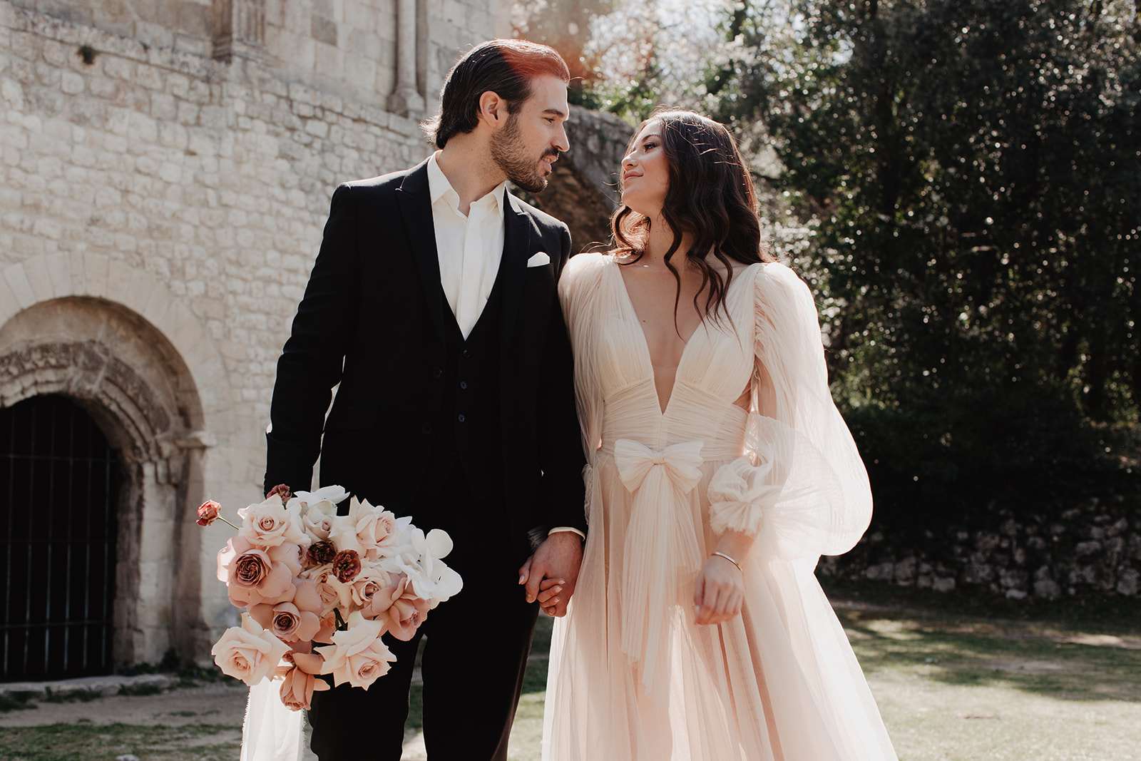 Bride in blush tulle gown with bow waist and mauve rose bouquet beside groom at stone abbey doorway