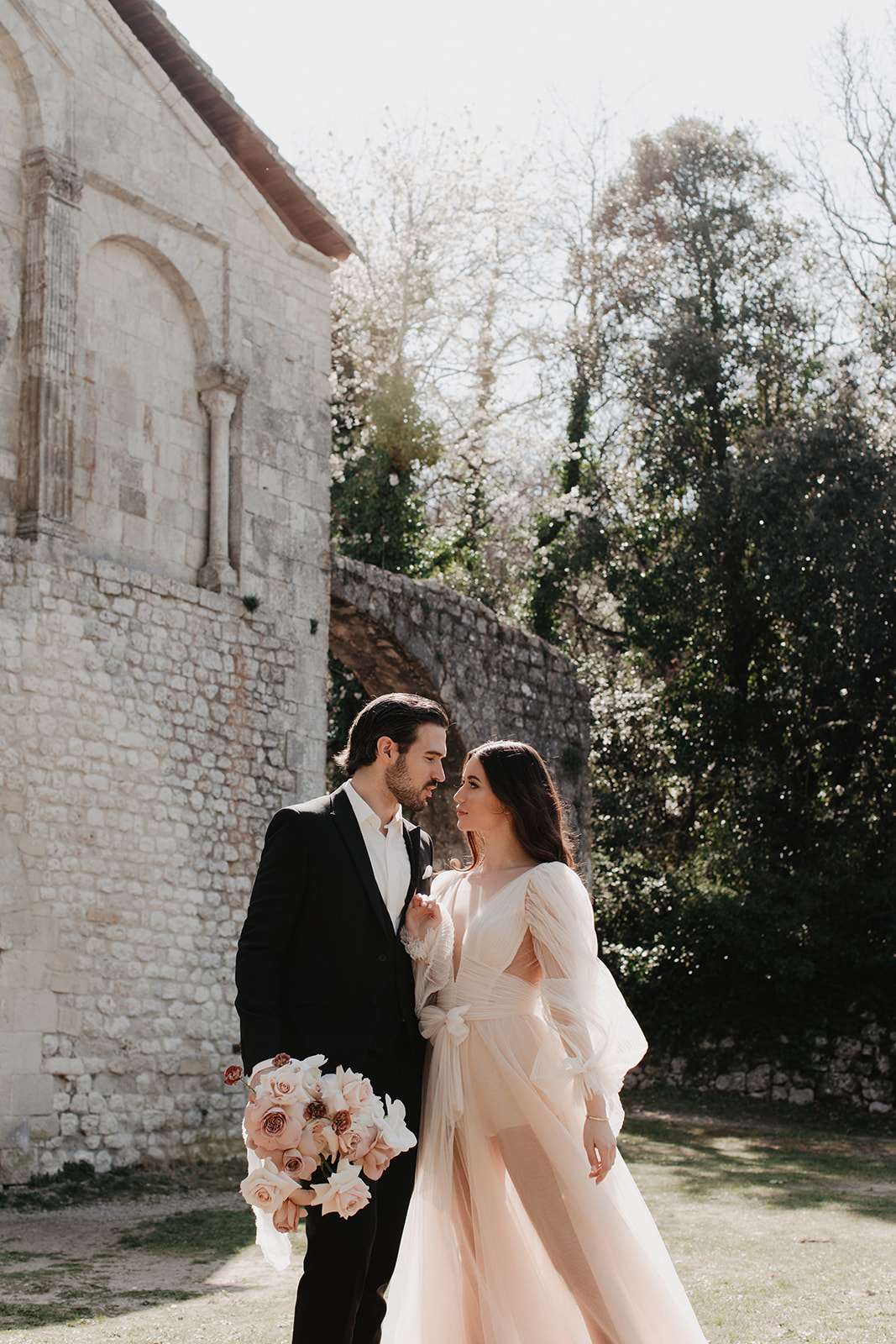 A couple portrait shot outdoors beside the stone facade of an ancient chapel or abbey, with historic carved stonework and arched architectural details visible. The groom wears a dark navy-black suit with a white shirt, and the bride wears a blush pink tulle gown with long sheer sleeves and a wrap-style bodice with a bow detail. She holds a lush round bouquet composed of blush and antique mauve garden roses with white accents. The two stand close together, faces nearly touching, looking at one another in a medium portrait composition with the stone building and trees in the background. The styling leans toward a romantic, classic aesthetic with a soft, warm color palette of blush and ivory against the aged stone setting.