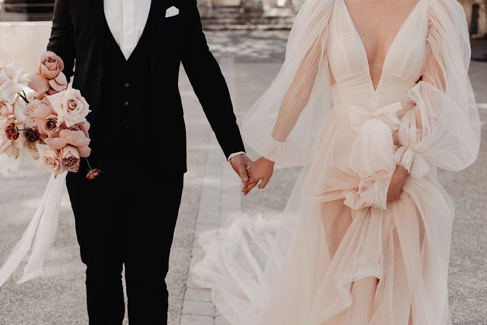Cropped portrait of couple holding hands showing blush chiffon gown and trailing rose bouquet with ribbons