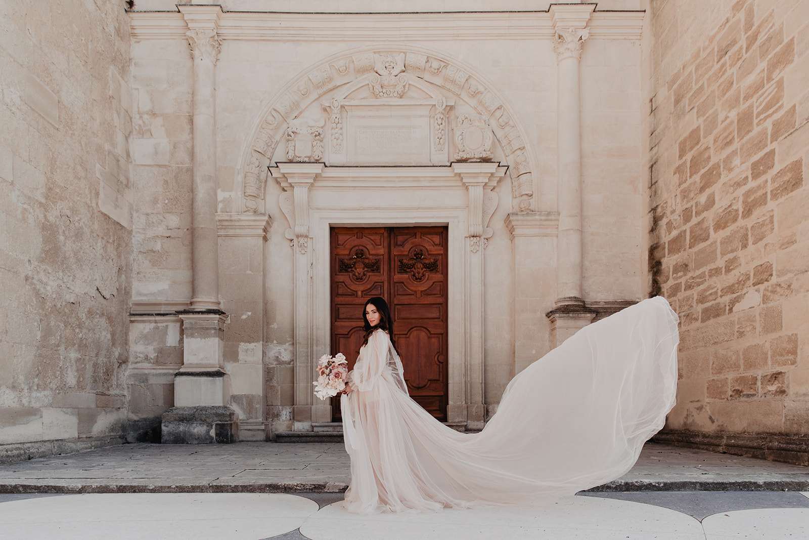 A bridal portrait shot outdoors in front of a historic French stone building featuring an ornately carved arched doorway with a large carved wooden double door. The bride, a dark-haired woman, stands centered in front of the doorway wearing a blush-to-ivory long-sleeved tulle gown with a dramatically billowing, wind-caught skirt and train extending across the right side of the frame. She holds a loose, garden-style bouquet in blush, dusty rose, and ivory tones, including what appear to be garden roses and ranunculus. The styling aesthetic is romantic and airy, with the soft blush palette of the dress complementing the warm cream tones of the carved stone architecture. The composition is a full-length environmental portrait with the architectural doorway framing the subject centrally.