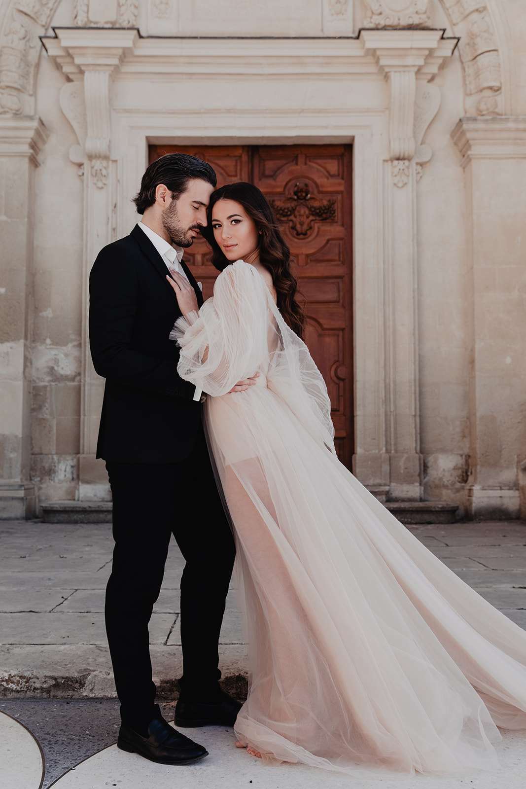 Bride in blush tulle gown with sheer sleeves and groom in black suit before carved stone doorway