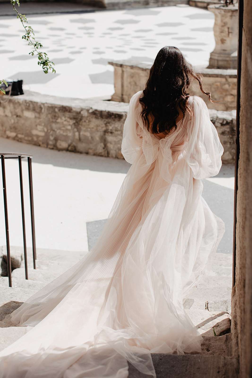 Bride from behind in blush tulle gown with puff sleeves and bow descending stone steps at chateau courtyard