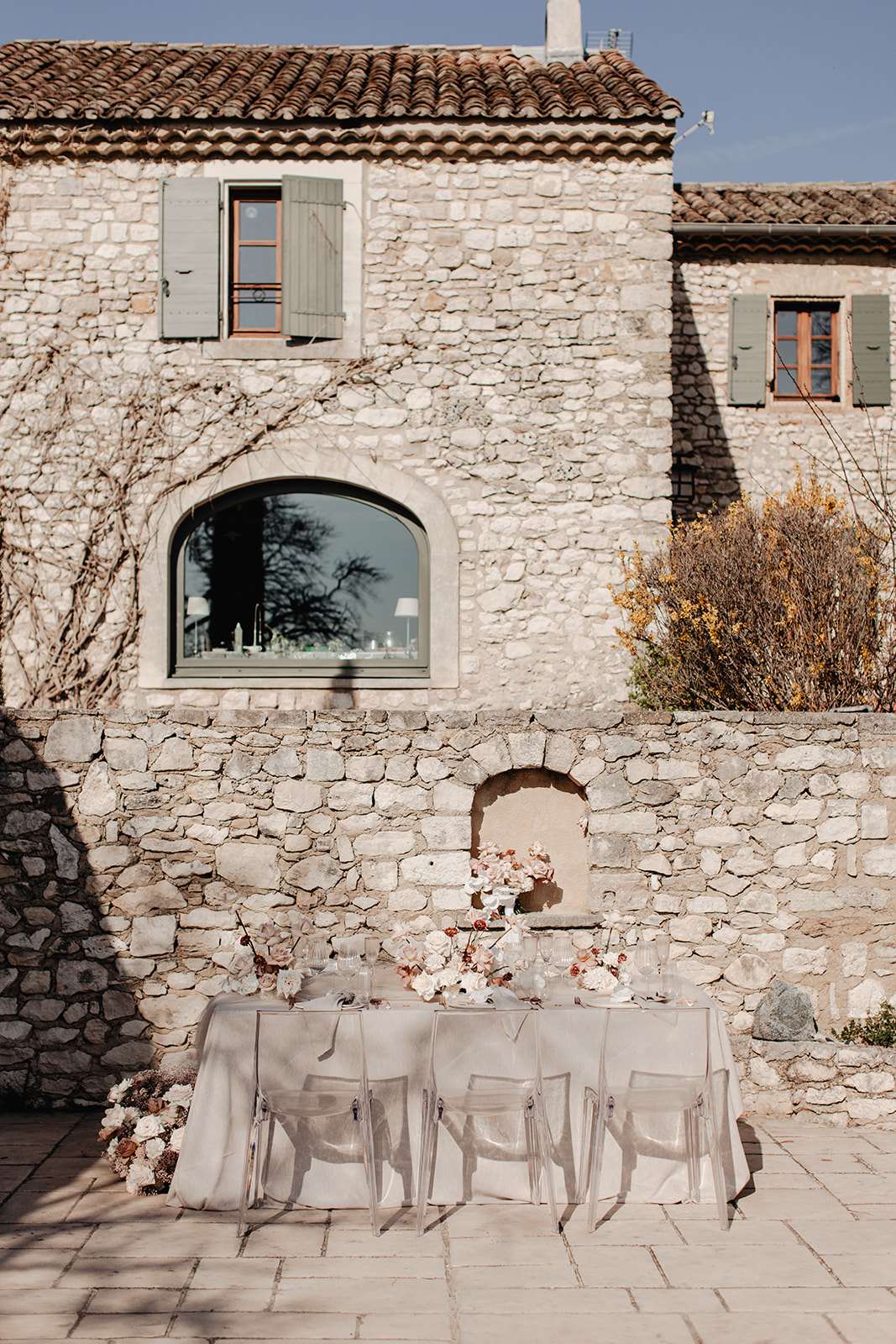 Ghost chairs at table with dusty rose and terracotta floral runner before Provencal stone farmhouse