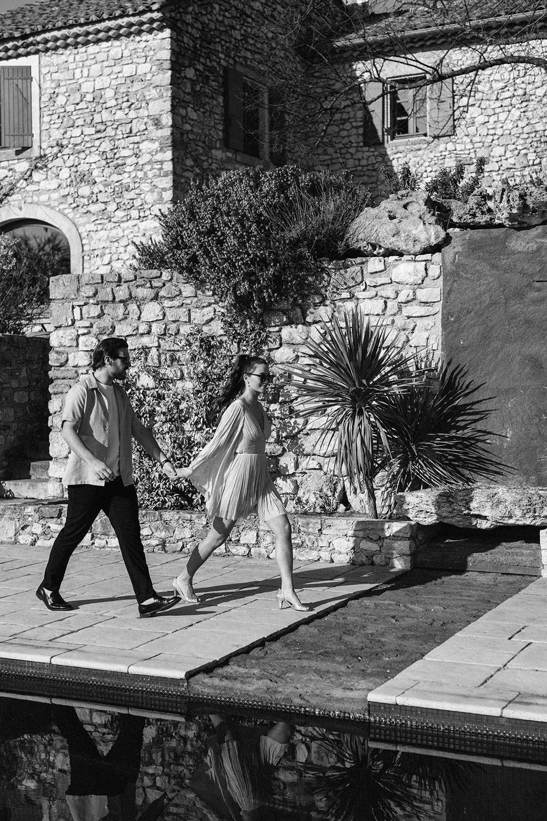 This black-and-white portrait shows a couple walking hand-in-hand along a stone pool surround at an outdoor venue. The woman wears a light-toned pleated midi skirt with a cropped long-sleeve top and heeled sandals, paired with sunglasses; the man wears dark trousers, a short-sleeve casual shirt, and loafers, also wearing sunglasses. The background features a rustic stone-built building with arched doorways, wooden shutters, and a large spiky yucca plant, characteristic of a southern French mas or domaine. The image is taken as a full-length candid walking shot with strong contrast between bright sunlight and deep shadows, with the couple's reflections visible in the pool water below.