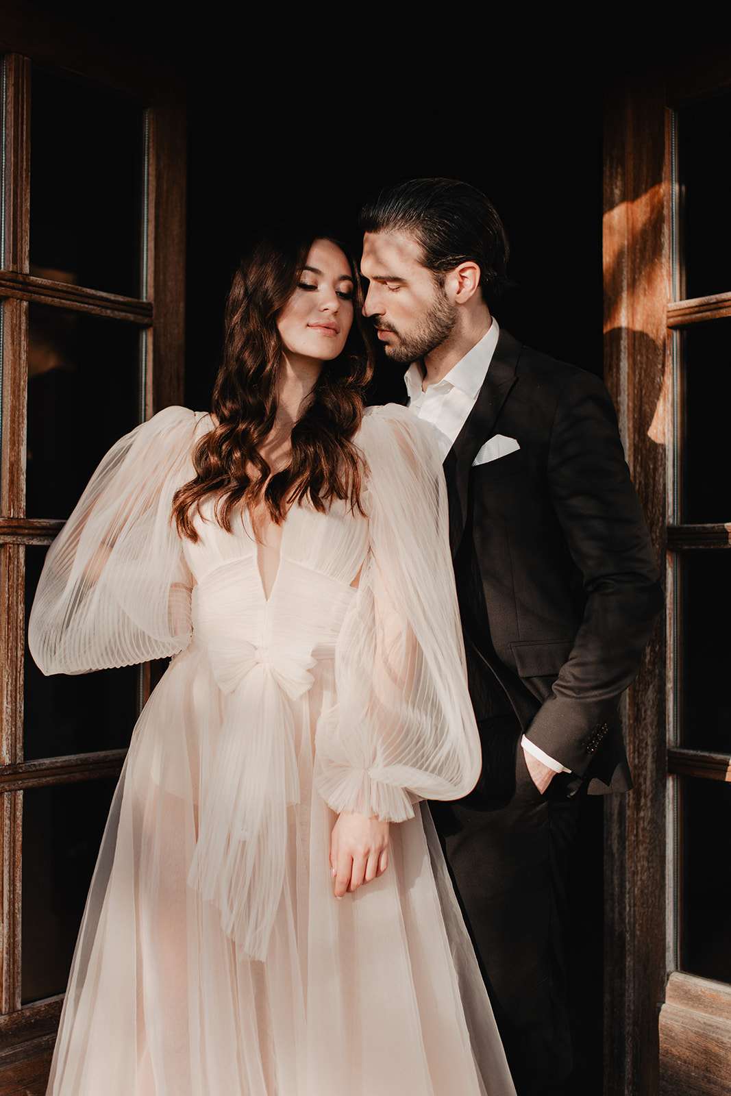 Moody portrait of couple before wooden doors with bride in pleated bishop-sleeve tulle gown
