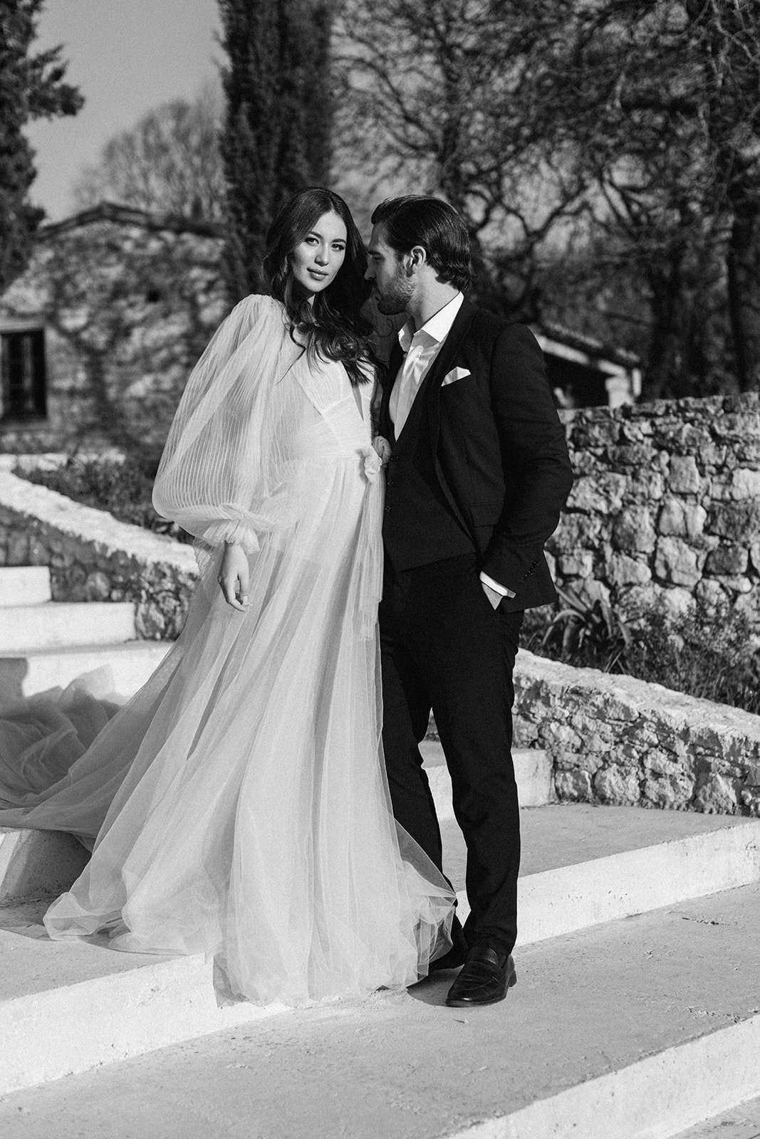 A black-and-white couple portrait taken outdoors on stone steps, with high contrast and bright highlights. The bride wears a flowing tulle gown with voluminous pleated bishop sleeves, a deep V-neckline, and a full skirt that billows slightly; the groom is dressed in a dark slim-fit suit with a white dress shirt, no tie, and a white pocket square. The bride looks toward the camera while the groom leans close to her, facing her profile in an intimate pose. The setting appears to be the grounds of a French stone-building property, with low terraced walls, cypress trees, and bare deciduous trees visible in the background, suggesting an off-season or early spring shoot.