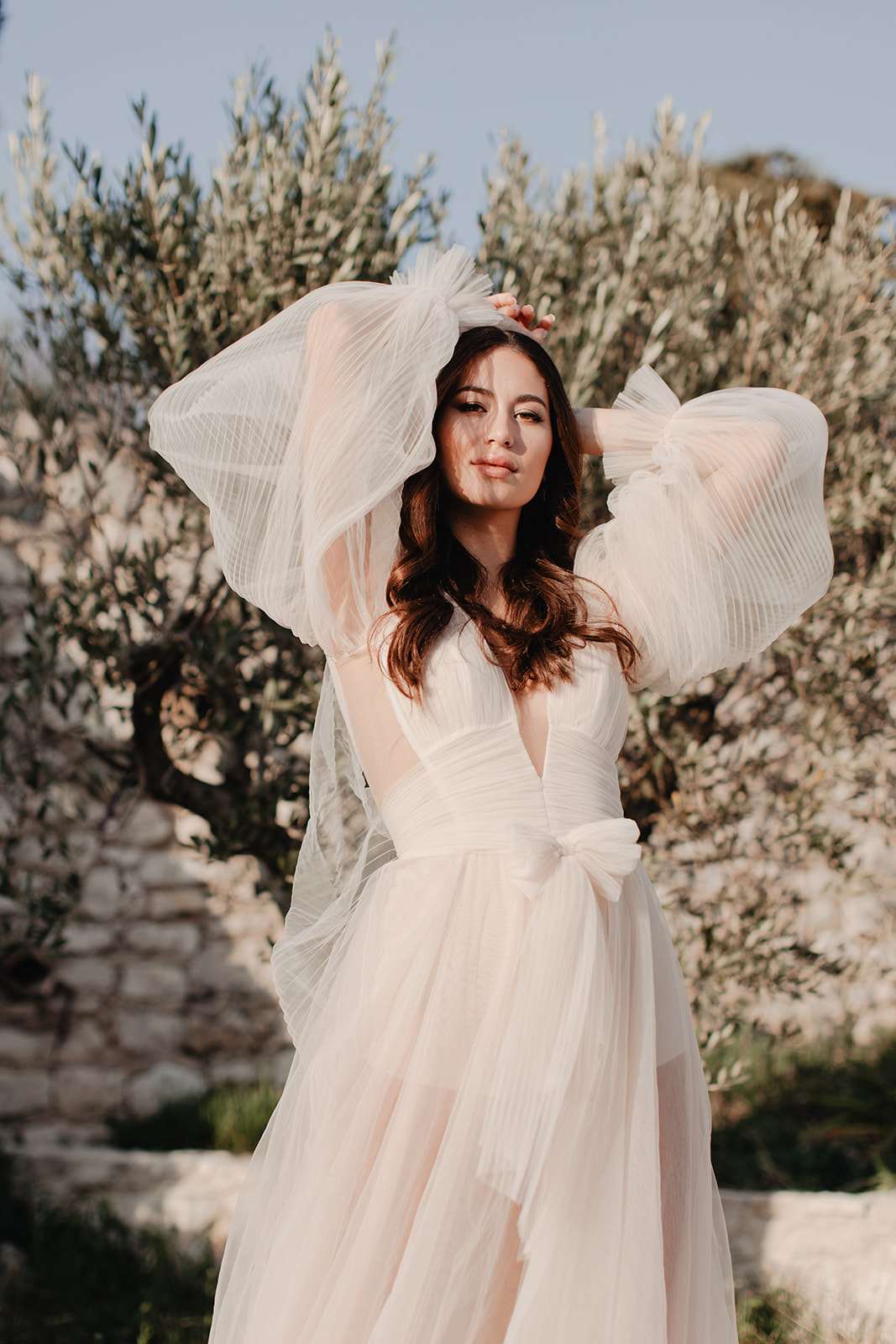 Bride in blush tulle gown with dramatic pleated sleeves posing among olive trees and dry stone wall
