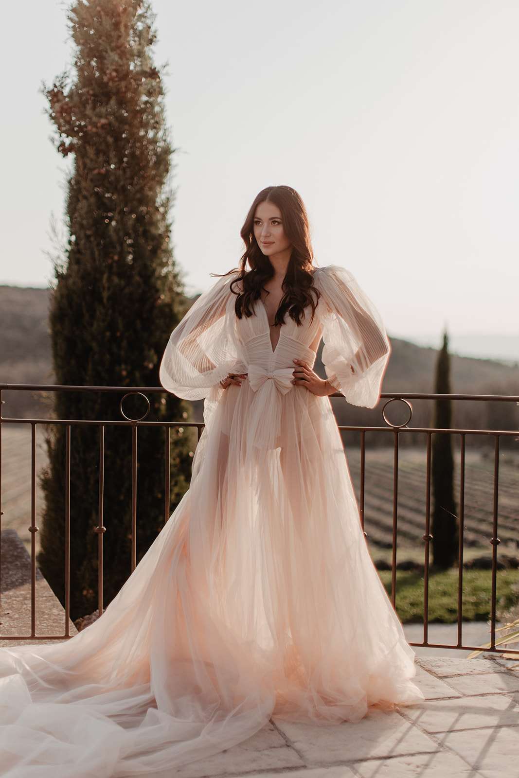 A bridal portrait of a single woman standing on an outdoor stone terrace with a wrought-iron railing, set against a backdrop of cypress trees and what appears to be a vineyard or agricultural landscape in the distance. The bride wears a blush-pink tulle ball gown with dramatic oversized bishop sleeves, a deep V-neckline, a structured bow detail at the waist, and a full sweeping train. Her dark hair is worn in loose waves, and she poses with her hands resting lightly on her waist, looking slightly off-camera. The image is a full-length portrait shot in warm golden-hour light, giving the scene a soft amber tone. The styling is romantic and fashion-forward, with the voluminous sleeves and pale blush palette as the clear focal point.