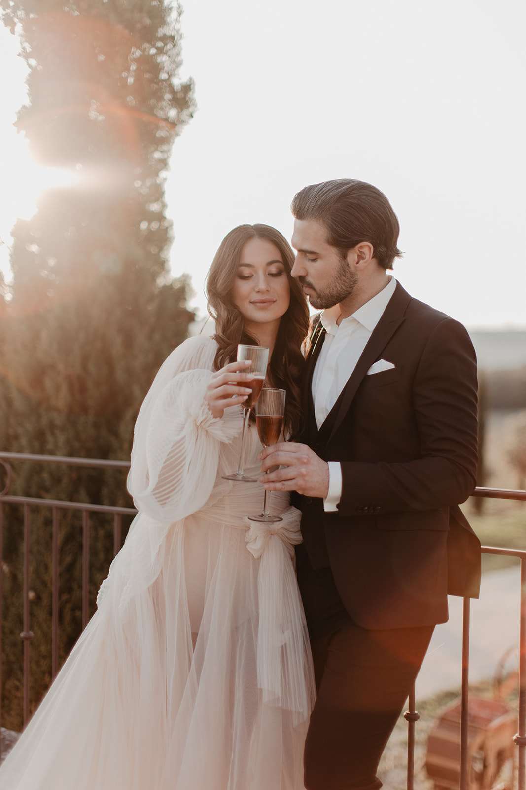 A couple portrait taken outdoors on a terrace or balcony with wrought iron railing, shot during golden hour with strong sun flare visible in the background. The bride wears a blush-toned tulle gown with voluminous pleated long sleeves and a sash bow at the waist, and has loose dark wavy hair; the groom wears a dark navy or black suit with a white dress shirt and white pocket square. Both hold ribbed champagne flutes containing what appears to be rosé. The groom leans his head toward the bride, who looks downward with eyes half-closed. The composition is a medium portrait shot with warm golden backlight creating a soft, hazy atmosphere around the couple.