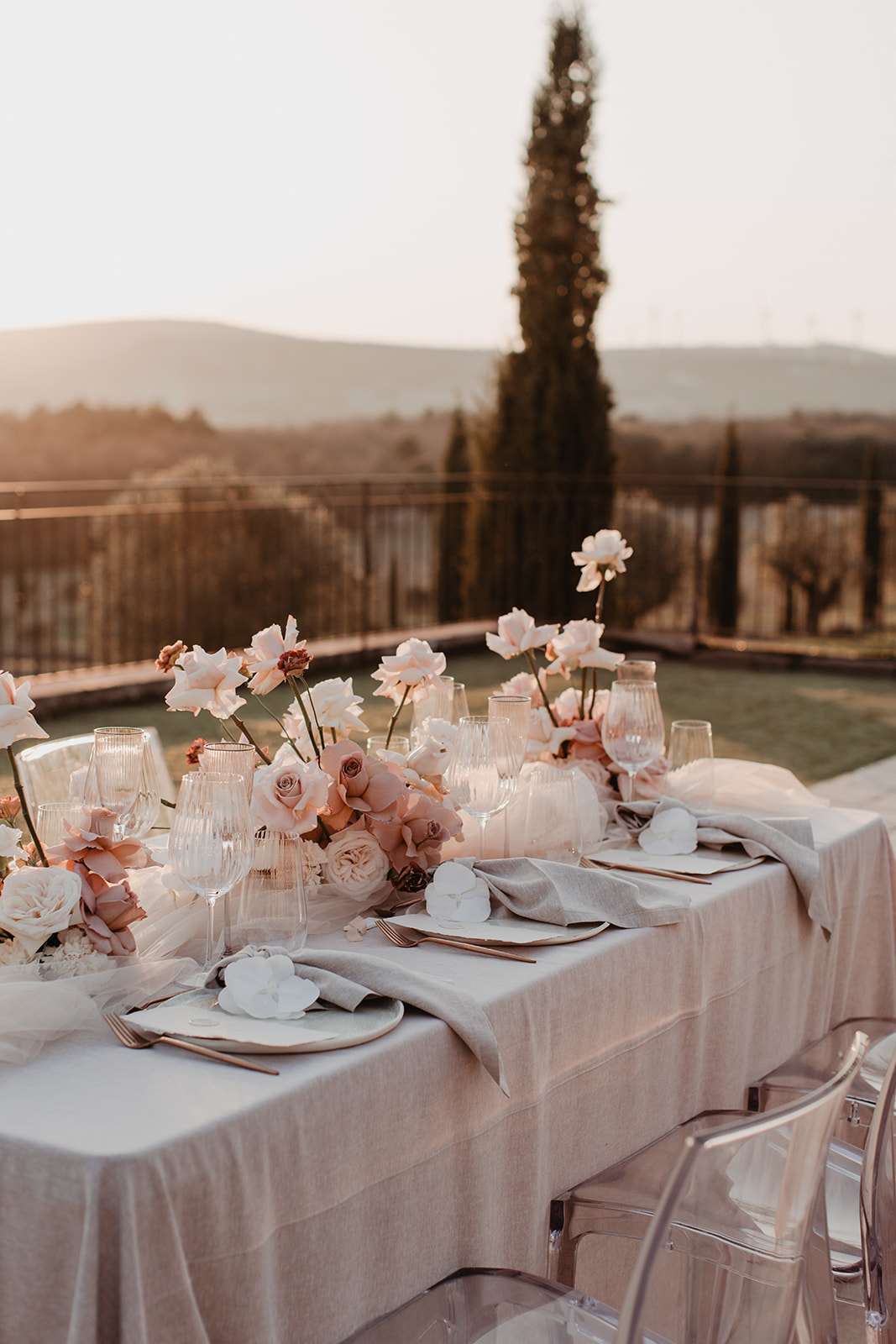 Dusty rose and terracotta garden roses with white pillar candles and ghost chairs on terrace at golden hour