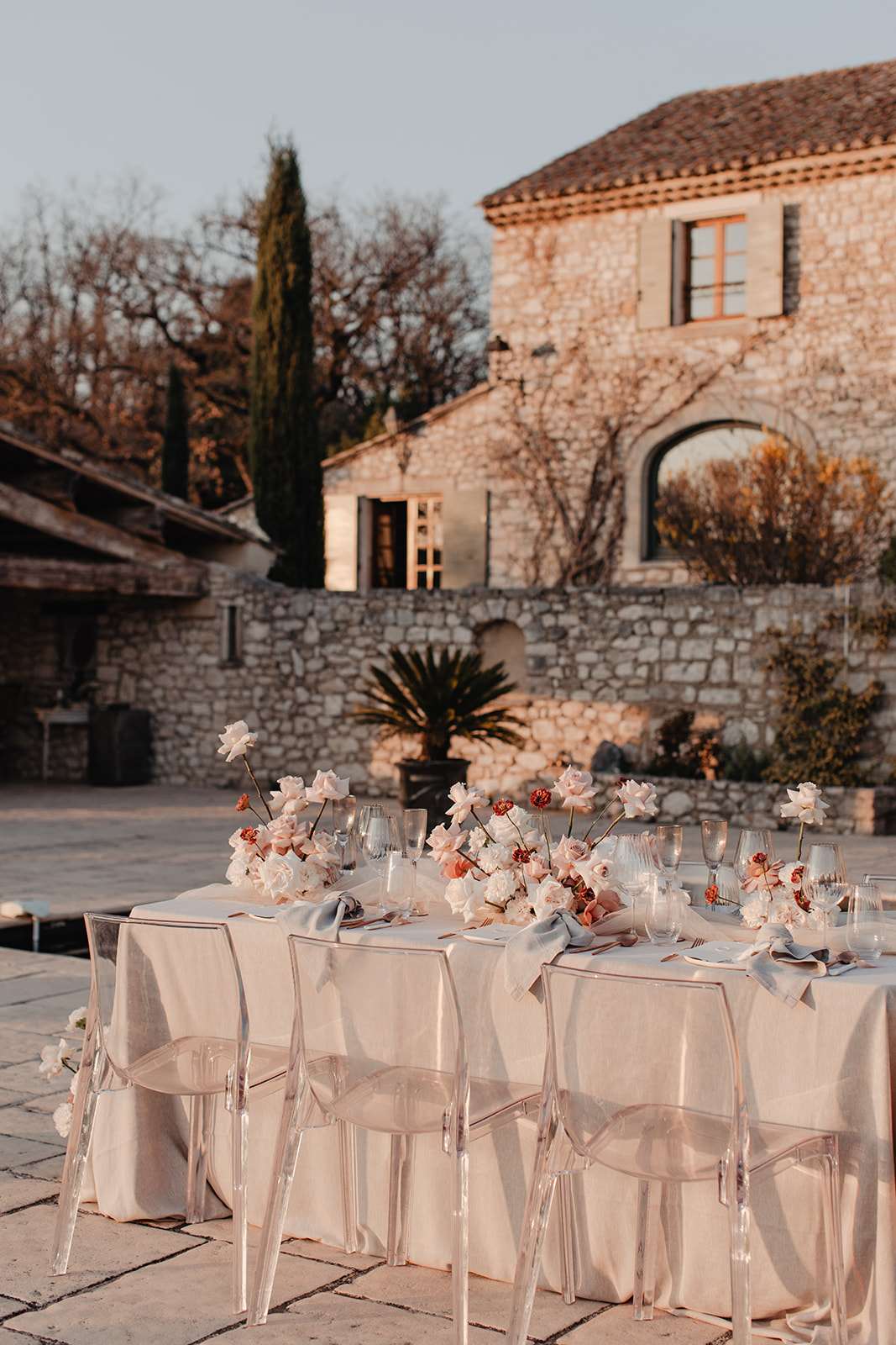 An outdoor reception table is set in the courtyard of a Provençal stone mas, photographed at golden hour from a slight elevated angle. The long rectangular table is dressed in a cream linen tablecloth with slate-blue linen napkins, clear acrylic ghost chairs, and glassware including wine glasses and champagne flutes. The floral centerpiece runs the length of the table and features blush garden roses, ivory peonies, and terracotta-toned ranunculus on tall single stems mixed with low clustered blooms, creating an organic, modern arrangement. The decor palette combines cream, blush, dusty blue, and warm terracotta against the backdrop of the rustic stone venue architecture. Potential venue feature image.