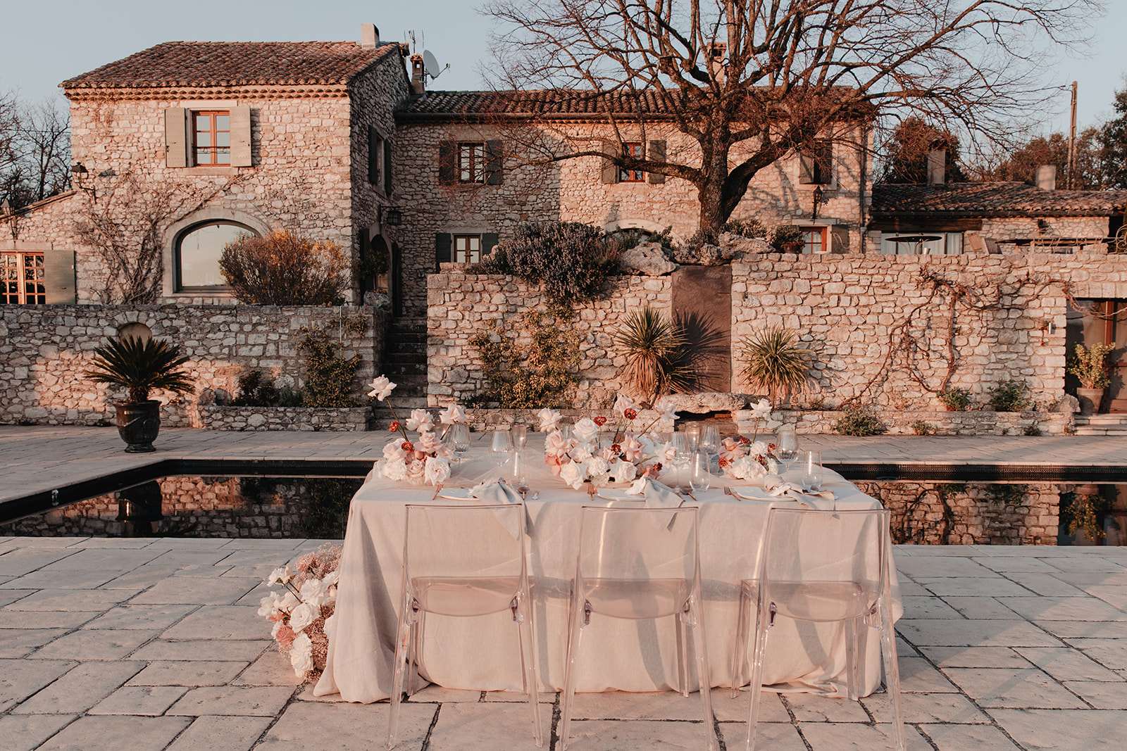 An outdoor wedding reception tablescape styled on a stone-paved terrace beside a dark-tiled reflecting pool, with a multi-building Provençal stone mas in the background. The rectangular table is dressed in a floor-length ivory linen tablecloth and surrounded by clear acrylic ghost chairs. Florals in blush pink, white, and terracotta tones — including garden roses, ranunculus, and delicate branching stems — run along the center of the table and spill onto the ground at one corner. Place settings include clear glassware, white linen napkins, and white plates. The wide-angle shot captures both the table styling and the full venue facade, photographed in warm golden-hour light. Potential venue feature image.
