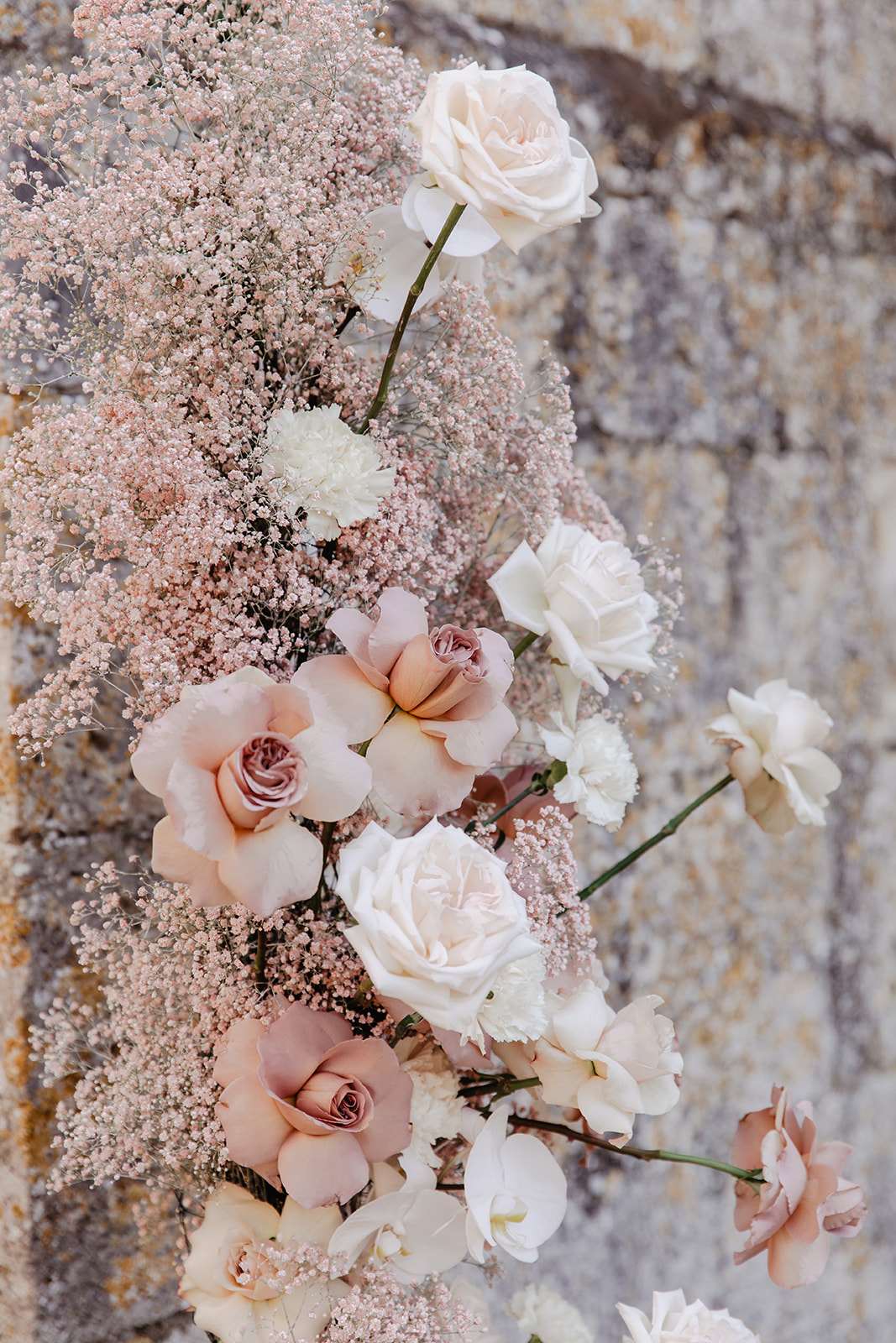 Floral arrangement of blush garden roses, mauve roses, ivory roses, white orchids, and baby's breath against stone wall