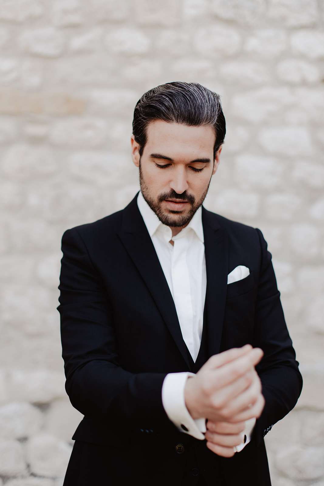 A groom getting ready portrait shot outdoors against a pale stone wall. The groom is adjusting his cufflinks, looking down at his wrists. He is wearing a navy black suit jacket, a white dress shirt with French cuffs, and a white pocket square. His dark hair is slicked back and he has a short beard. The composition is a close-up portrait with shallow depth of field, keeping the groom sharp against a softly blurred background. The overall styling is classic and formal.