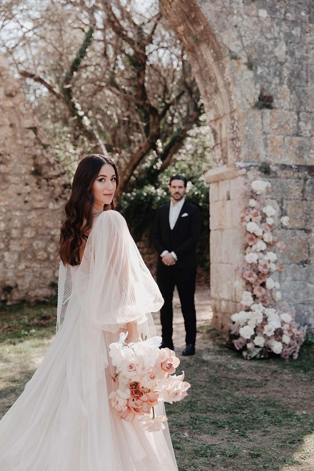 Bride holding blush and white bouquet looking over her shoulder with groom standing under a stone archway with floral inst...