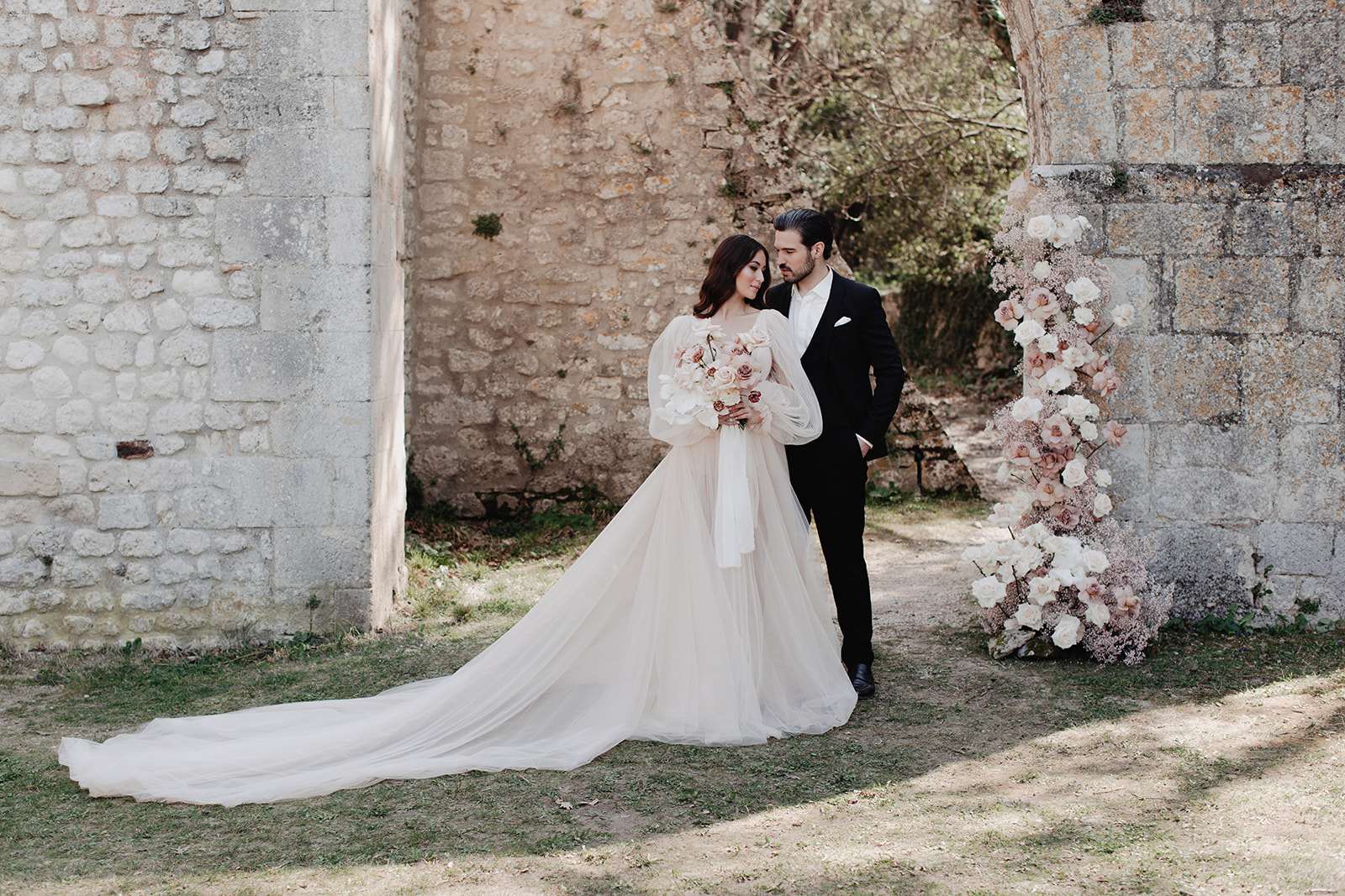 A couple portrait taken outdoors against the ruins of a historic stone building. The bride wears a blush tulle ballgown with voluminous long puffed sleeves and an extended cathedral-length train, and holds a bouquet of blush orchids, soft pink roses, and ivory blooms with trailing white ribbon. The groom is dressed in a black tuxedo with a white pocket square. They stand close together, faces nearly touching, in a quiet intimate pose. To the right, a tall floral column arrangement features white and blush roses, ranunculus, and baby's breath. The styling is classic-romantic with a soft blush and ivory color palette. The composition is a full-length wide portrait shot.