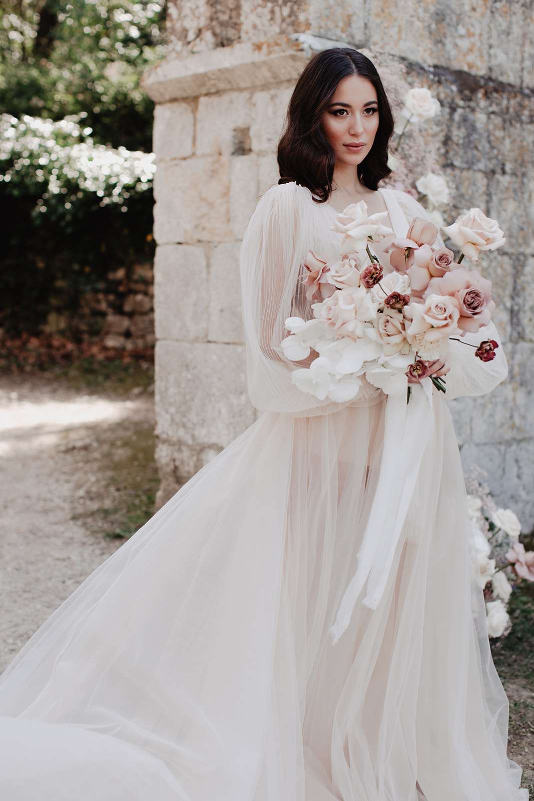 Bride in blush tulle gown with pleated sleeves holding bouquet of orchids, blush roses, and burgundy blooms