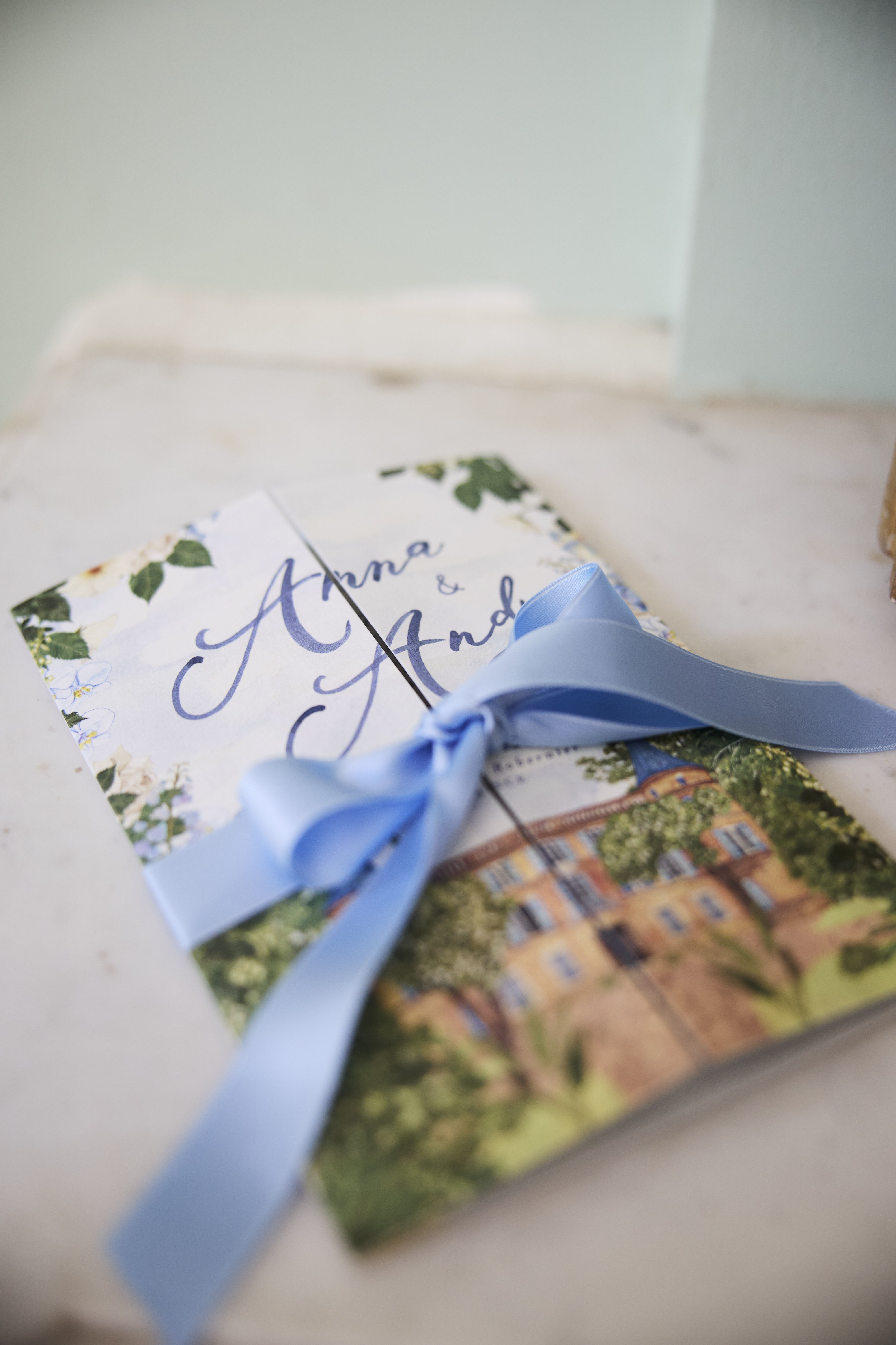 Close-up detail shot of wedding stationery resting on a marble surface. Two folded booklets are tied together with a powder blue satin ribbon bow — one features navy calligraphy script reading 'Anna & And...' (second name partially obscured) on a white background bordered with illustrated white florals and green foliage, while the other displays a watercolor illustration of a French château or villa surrounded by trees. The stationery palette combines navy blue, powder blue, white, and green, consistent with a classic garden-inspired wedding design.