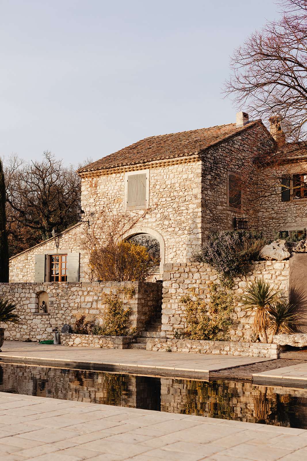 An exterior wide shot of a traditional Provençal stone farmhouse, likely a mas, photographed in warm natural light. The two-storey building features pale limestone construction, terracotta roof tiles, sage-green wooden shutters, and a characteristic arched stone gateway connecting two sections of the property. A rectangular pool or reflecting basin with dark tile edging sits in the foreground on a stone-paved terrace, with the building's façade mirrored in the still water. Stone retaining walls with planted shrubs and a yucca plant separate the pool terrace from the main structure. Potential venue feature image.