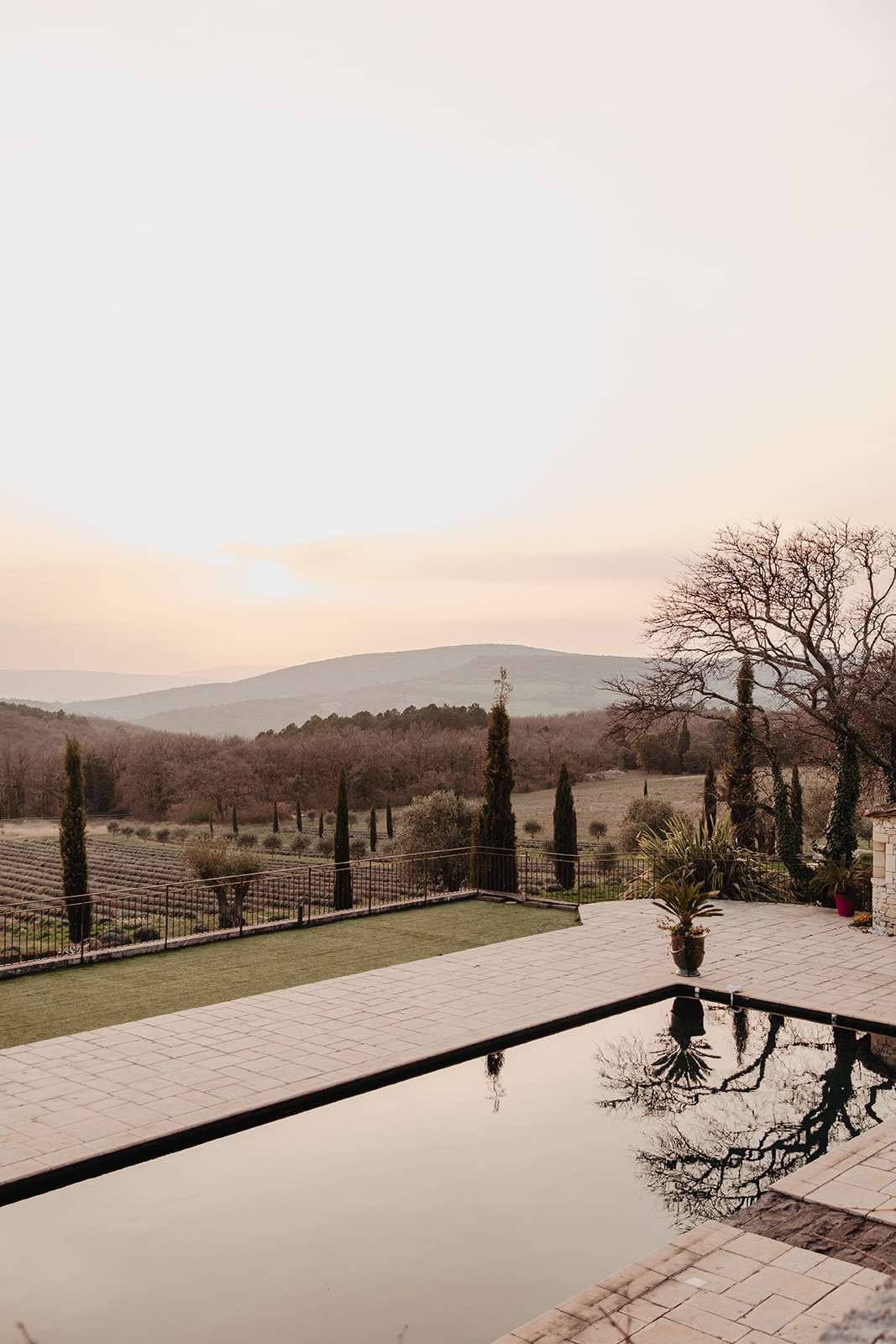 A wide exterior shot of a Provençal estate venue at dusk, with no people or wedding activity visible. The foreground features a dark-edged infinity or reflecting pool set into a pale stone terrace, with the still water mirroring surrounding trees. A manicured lawn area and a potted palm sit beside the pool, bordered by a wrought-iron railing. Beyond the grounds, rows of what appear to be lavender fields and cypress trees line the property, with rolling hills and a warm peach-toned horizon in the background. The overall palette is muted — warm stone, deep green cypress, and soft amber light. Potential venue feature image.