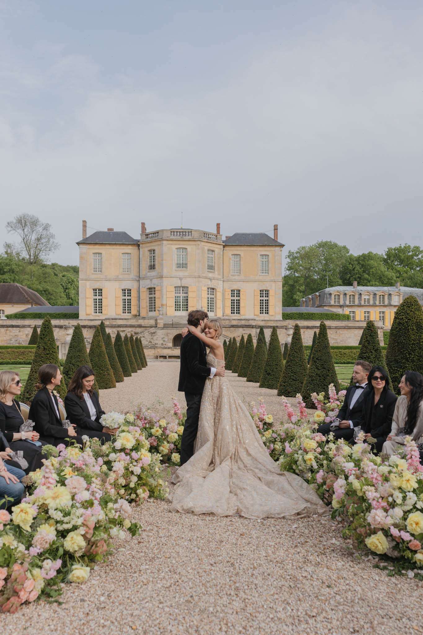 First kiss on chateau allee with blush and peach ground florals conical topiary and honey stone facade behind couple
