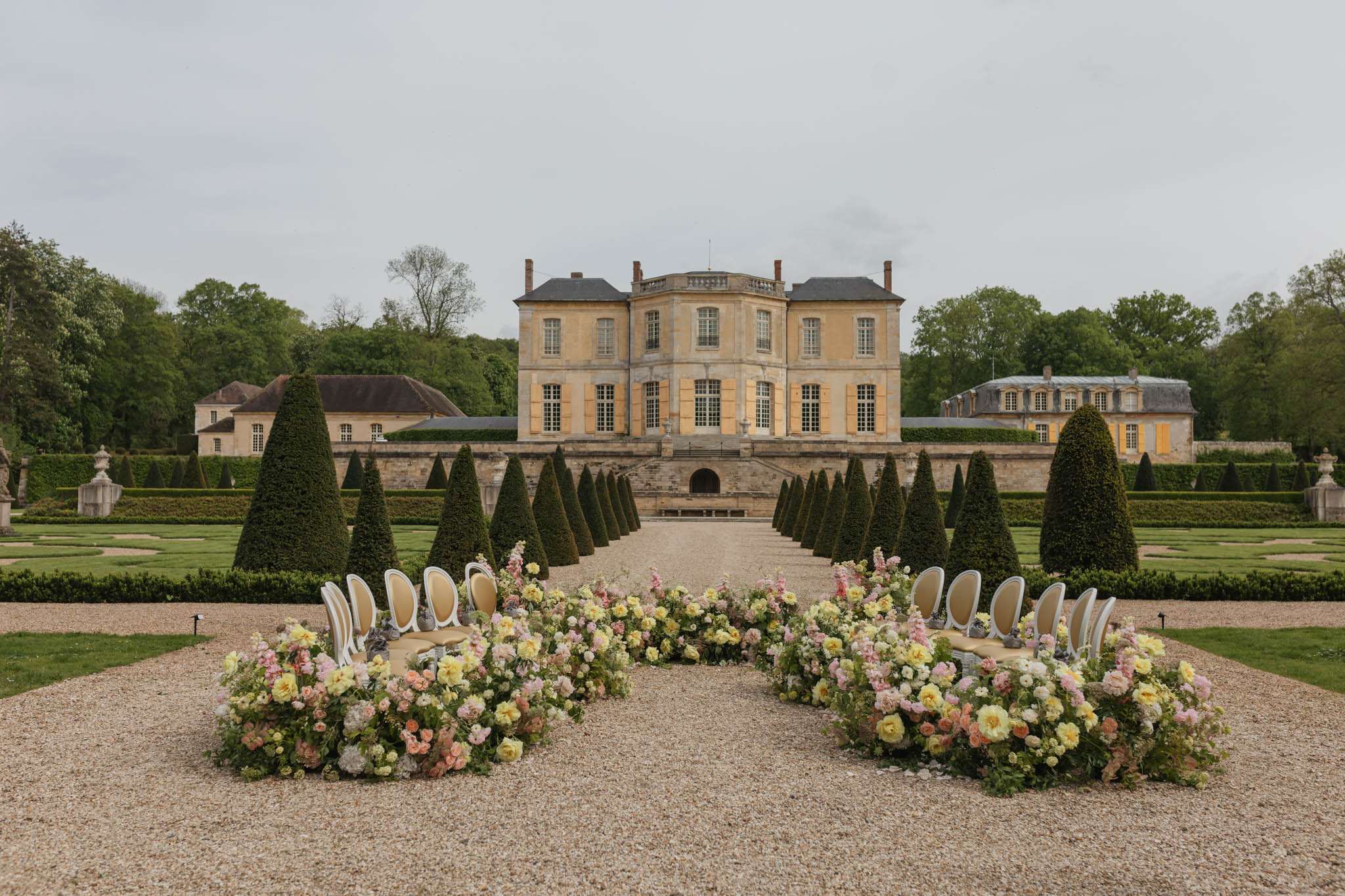 Ceremony setup on chateau parterre with yellow rose and pink peony installations and topiary allee