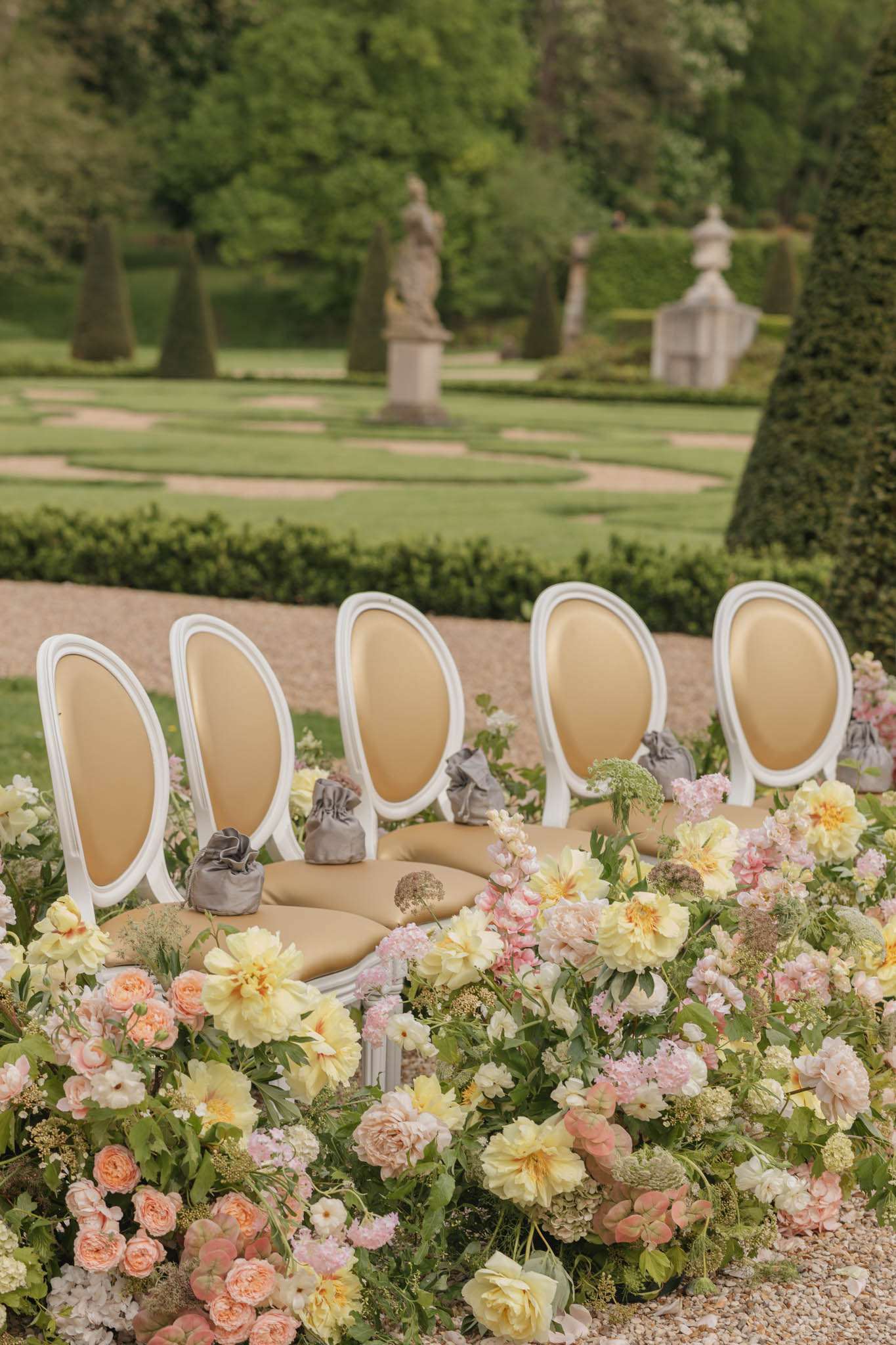 Ceremony chairs with velvet favor bags surrounded by yellow peony and peach rose ground floral installation in parterre ga...