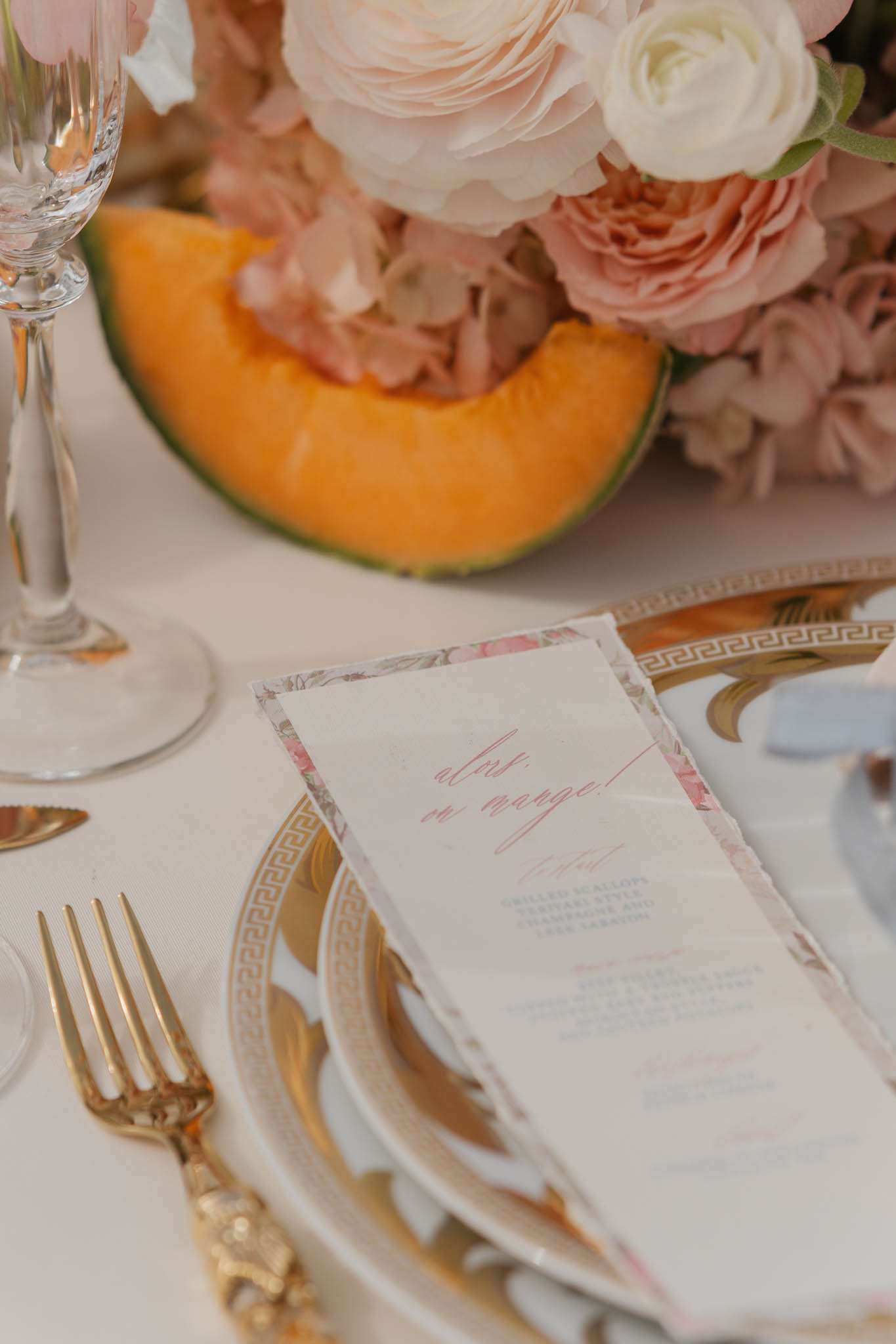 A close-up detail shot of a wedding reception place setting on a cream linen tablecloth. The place setting features a gold-rimmed plate with a Greek key border pattern, ornate gold flatware, and a crystal champagne flute. A printed menu card with a floral border and pink script reading "alors, on mange!" is placed on the plate. In the background, a slice of cantaloupe melon is visible alongside a floral centerpiece of blush garden roses, dusty pink hydrangeas, and ivory ranunculus. The overall decor palette is blush, ivory, and gold with a classic French-inspired aesthetic.