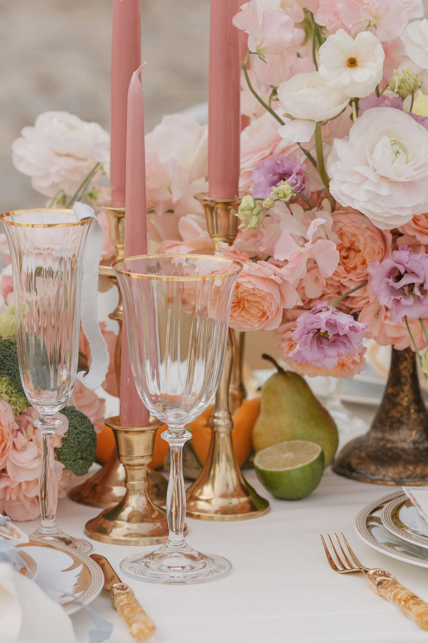 Maximalist tablescape with peach roses blush peonies dusty rose tapers gold cutlery crystal glassware and scattered fruit