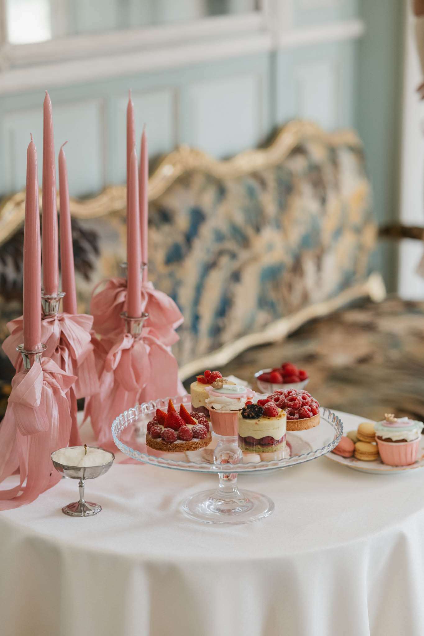 Wedding dessert table with raspberry tarts, macarons, and cupcakes beside dusty rose taper candles in a chateau salon