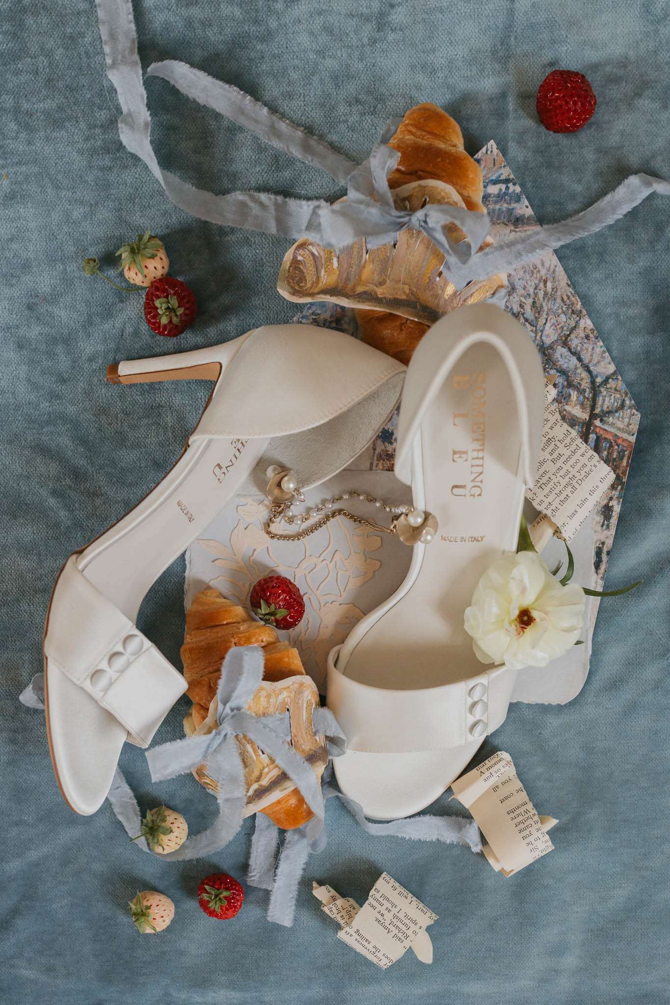 A flat lay detail shot of a pair of ivory bridal heels labeled 'Made in Italy' with pearl and gold chain anklet jewelry draped across them, arranged on a dusty blue linen textile. The styled composition includes golden croissants, fresh red strawberries, a cream peony bloom, torn strips of vintage book pages, a postcard featuring a city scene, and frayed pale blue silk ribbons scattered around the shoes. The shoes feature a peep-toe silhouette with pearl button detailing on the straps and a rose gold stiletto heel. The overall styling palette is ivory, dusty blue, and red with a whimsical French-inspired theme.