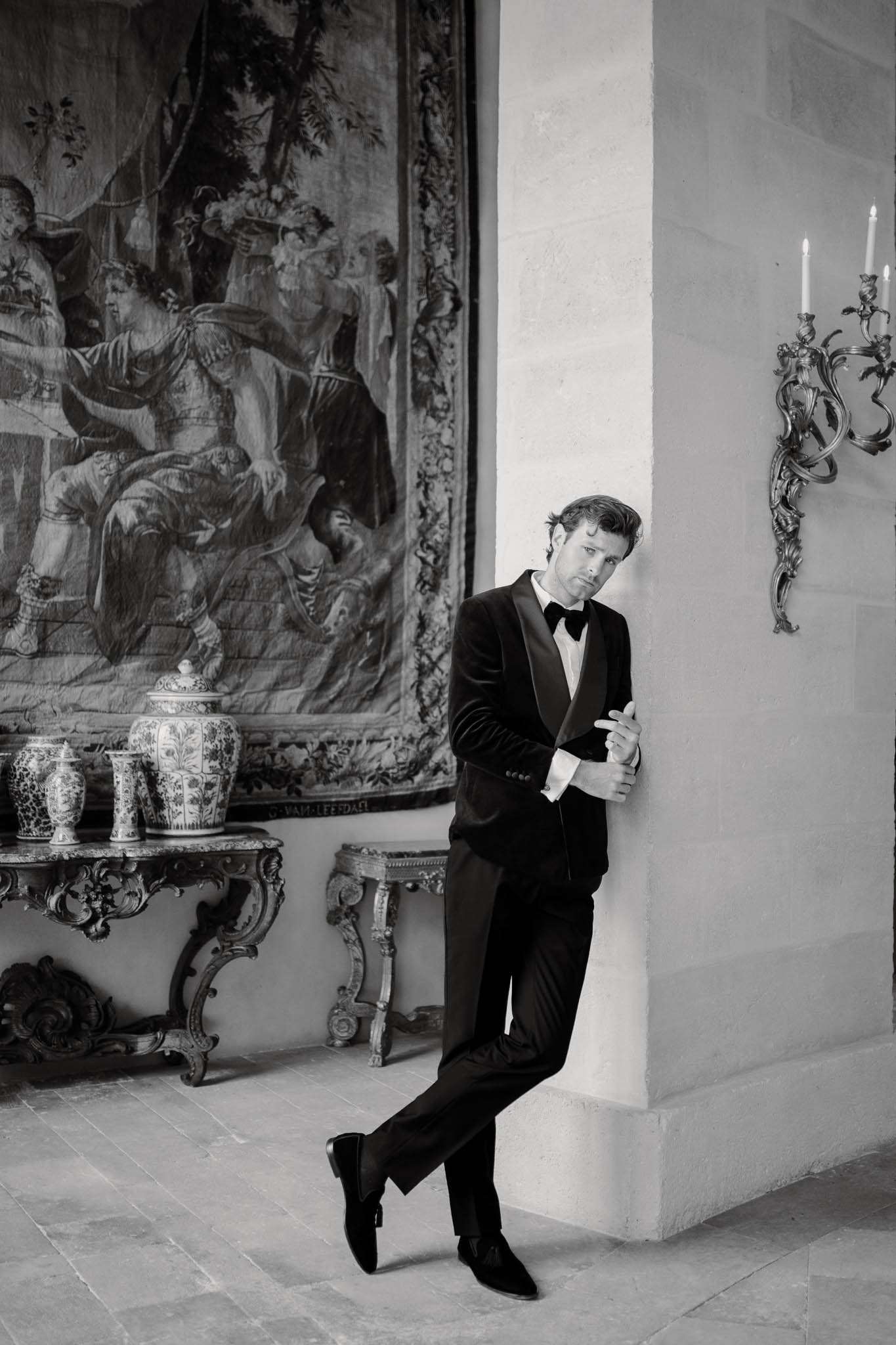 Black-and-white portrait of groom in velvet dinner jacket leaning against stone pillar inside French chateau