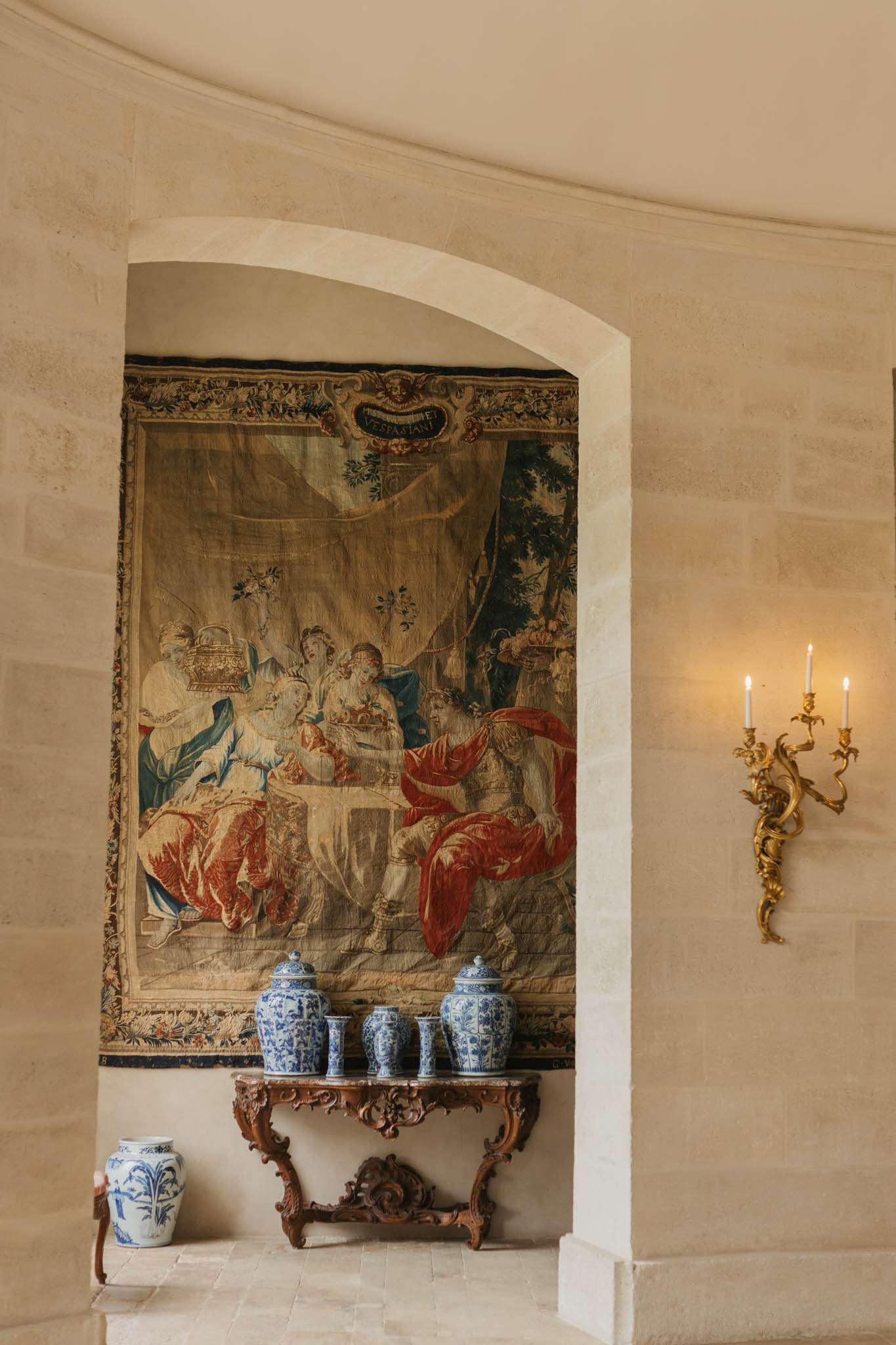 Stone corridor with Vespasiani tapestry, carved walnut console, blue-and-white porcelain, and gilt sconces