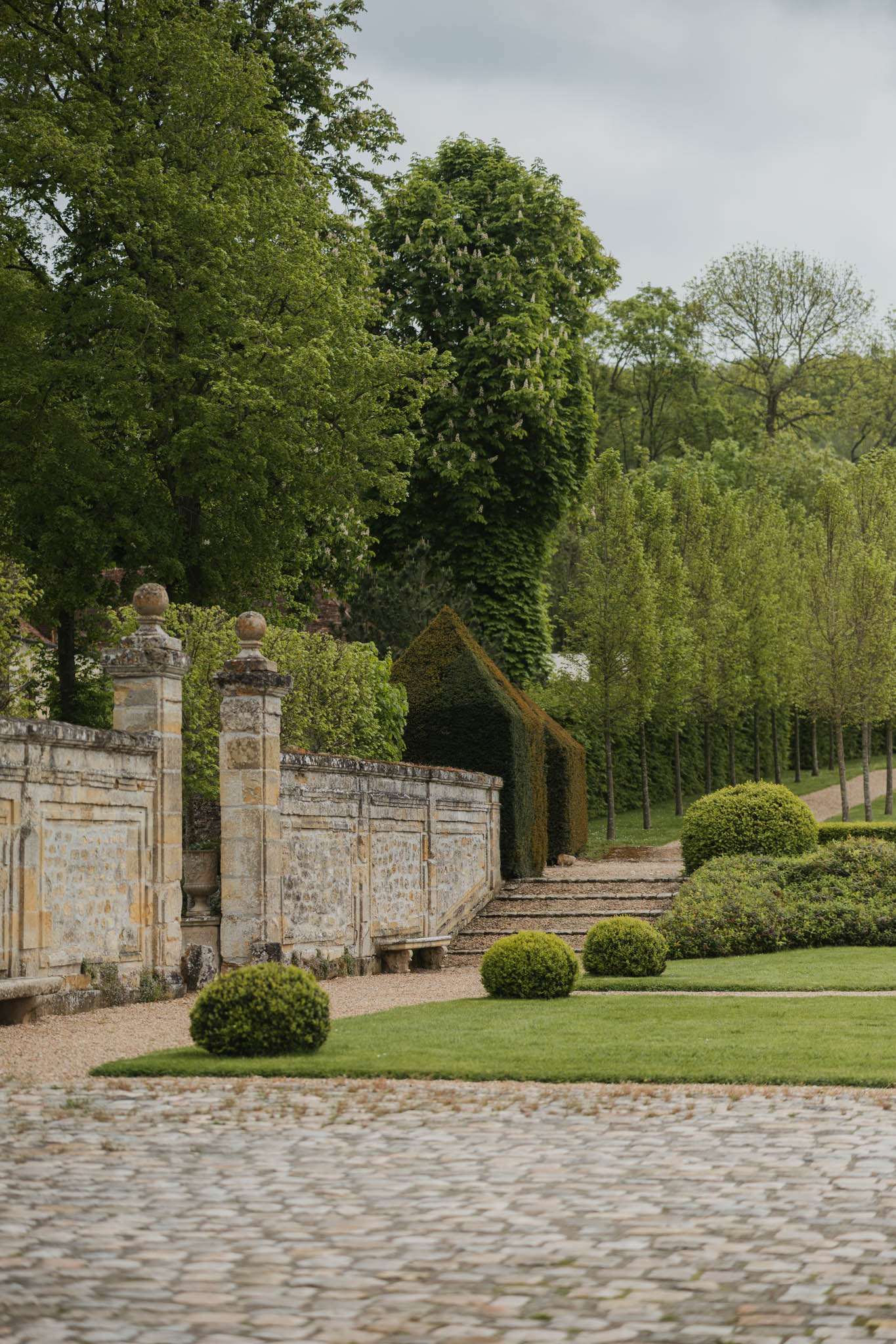 A venue grounds shot with no people present, showing the formal gardens and exterior walls of what appears to be a French château estate. The foreground features a cobblestone courtyard leading to a low stone perimeter wall with ornamental stone pillars topped with ball finials. Neatly clipped round boxwood topiaries are arranged along a gravel path, with manicured lawn strips between them. A hedged archway leads to a tree-lined avenue in the background. The composition is a medium wide shot taken from ground level along the wall. Potential venue feature image.
