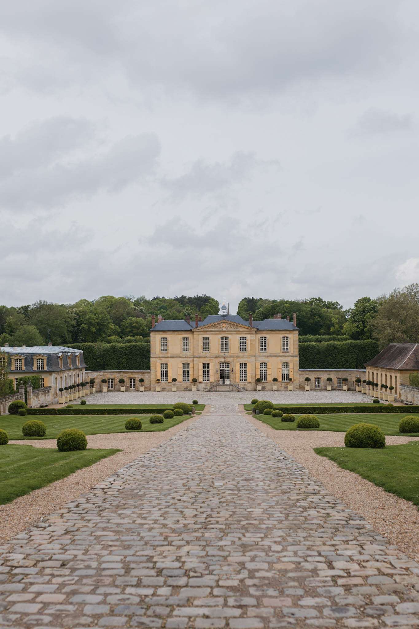 Symmetrical view of honey-limestone French chateau with slate mansard roof, formal parterre gardens, and cobblestone path
