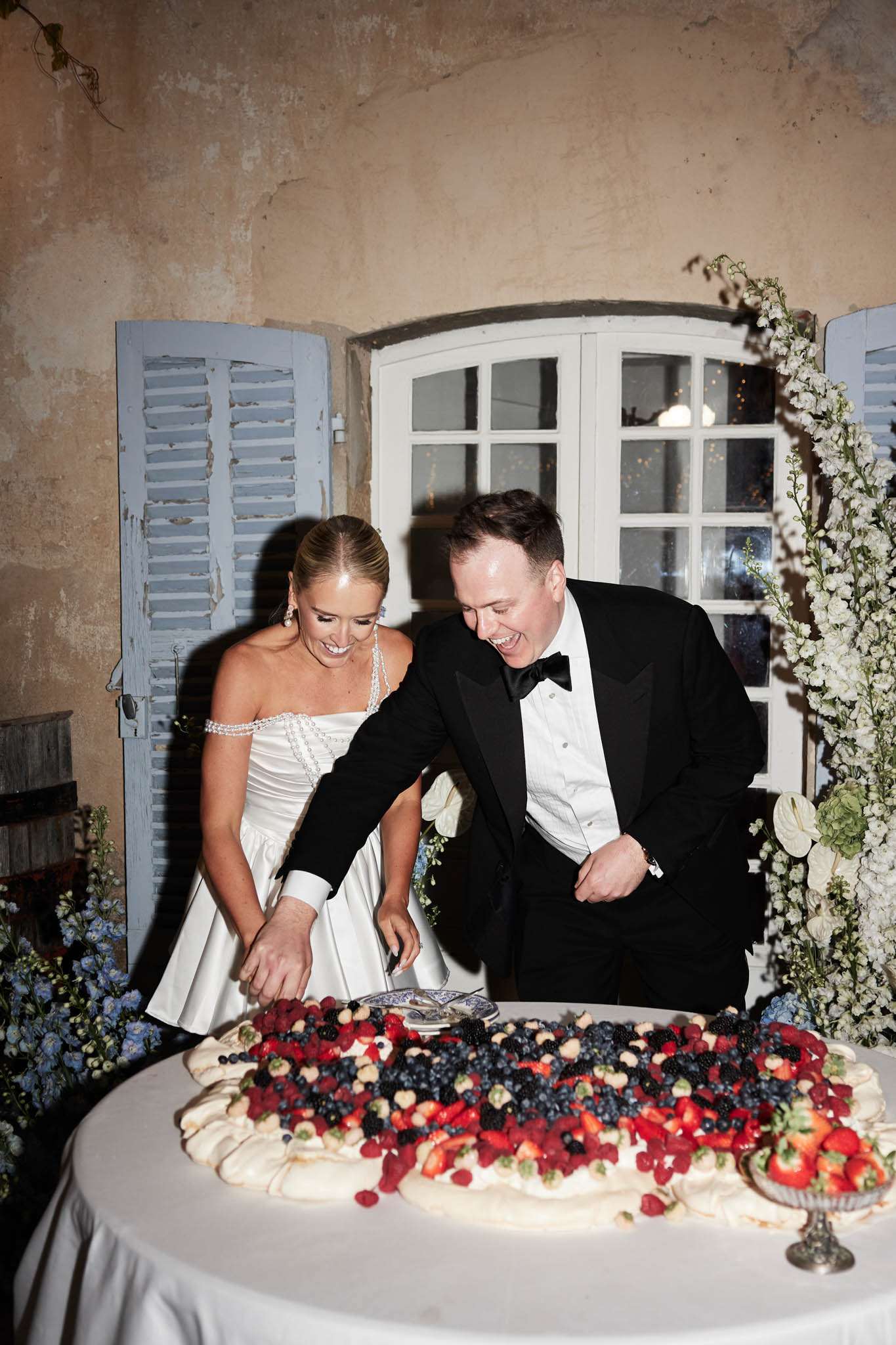Bride in white satin dress and groom in tuxedo cutting berry pavlova beside blue hydrangea arrangements