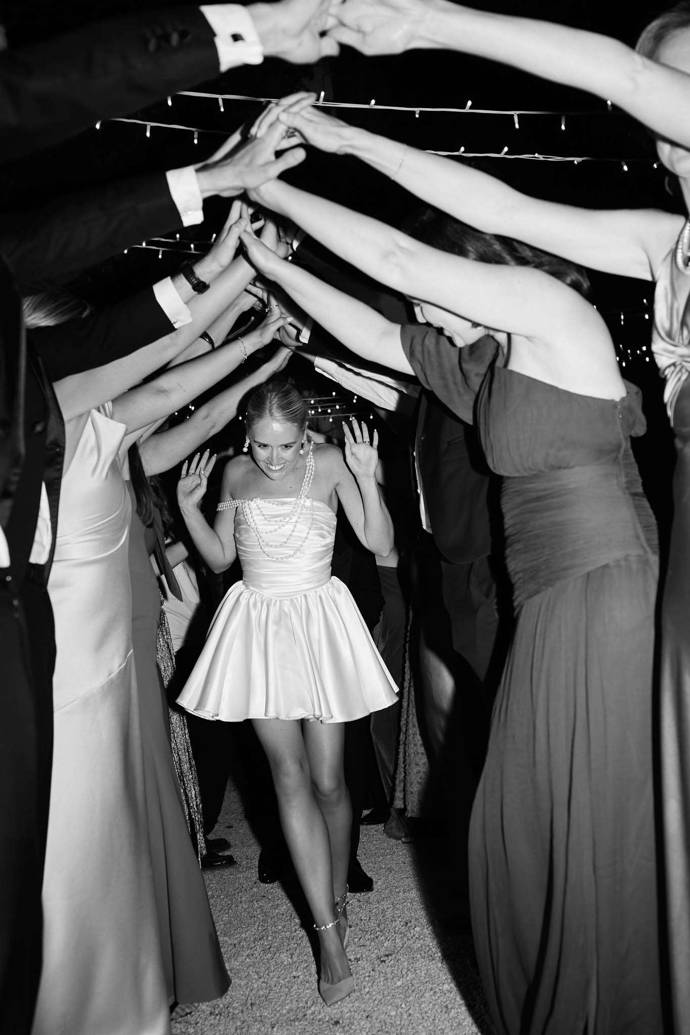 This black-and-white image captures a bride walking through a tunnel of raised arms formed by wedding guests during what appears to be a reception send-off or celebratory moment. The bride is wearing a short, strapless satin mini dress with a full skirt and layered pearl necklaces, accessorized with heeled sandals and an anklet. She is smiling broadly and raising her hands as she passes through the arch of arms. Guests on either side are dressed in formal attire — long floor-length gowns are visible on the right side, and a suit with French-cuff shirt is visible on the left. Fairy lights strung overhead are faintly visible in the dark background, suggesting an outdoor or covered evening setting. The image is shot from a low, centered perspective, framing the bride as the focal point of the composition.