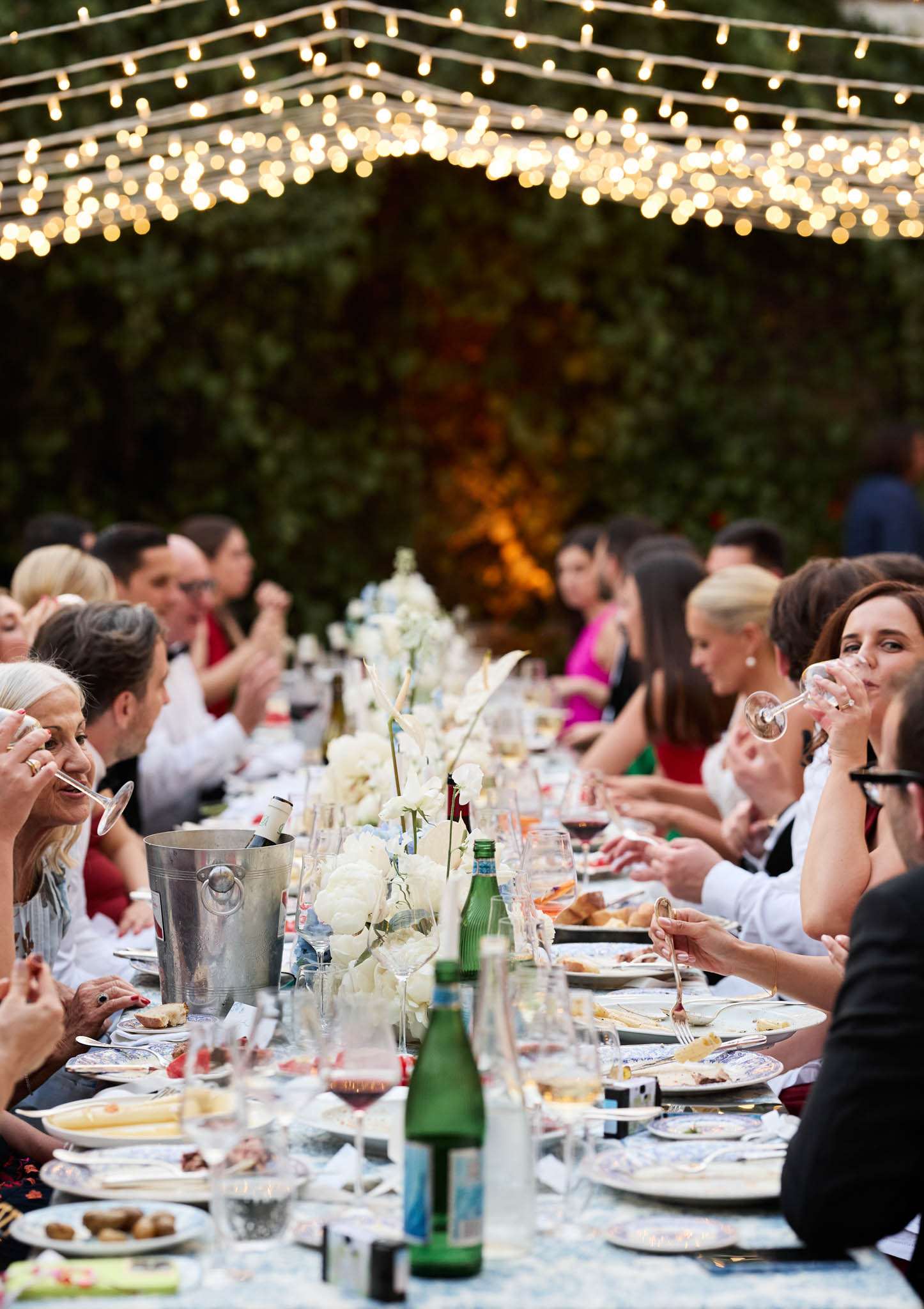 Twenty-five guests at banquet table under bistro lights beside ivy wall with white hydrangea and orchid runner