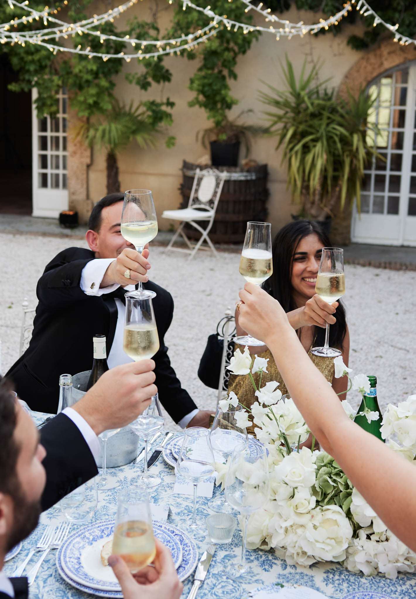 Guests raising champagne glasses at toile-dressed table with white roses sage hydrangeas and fairy lights