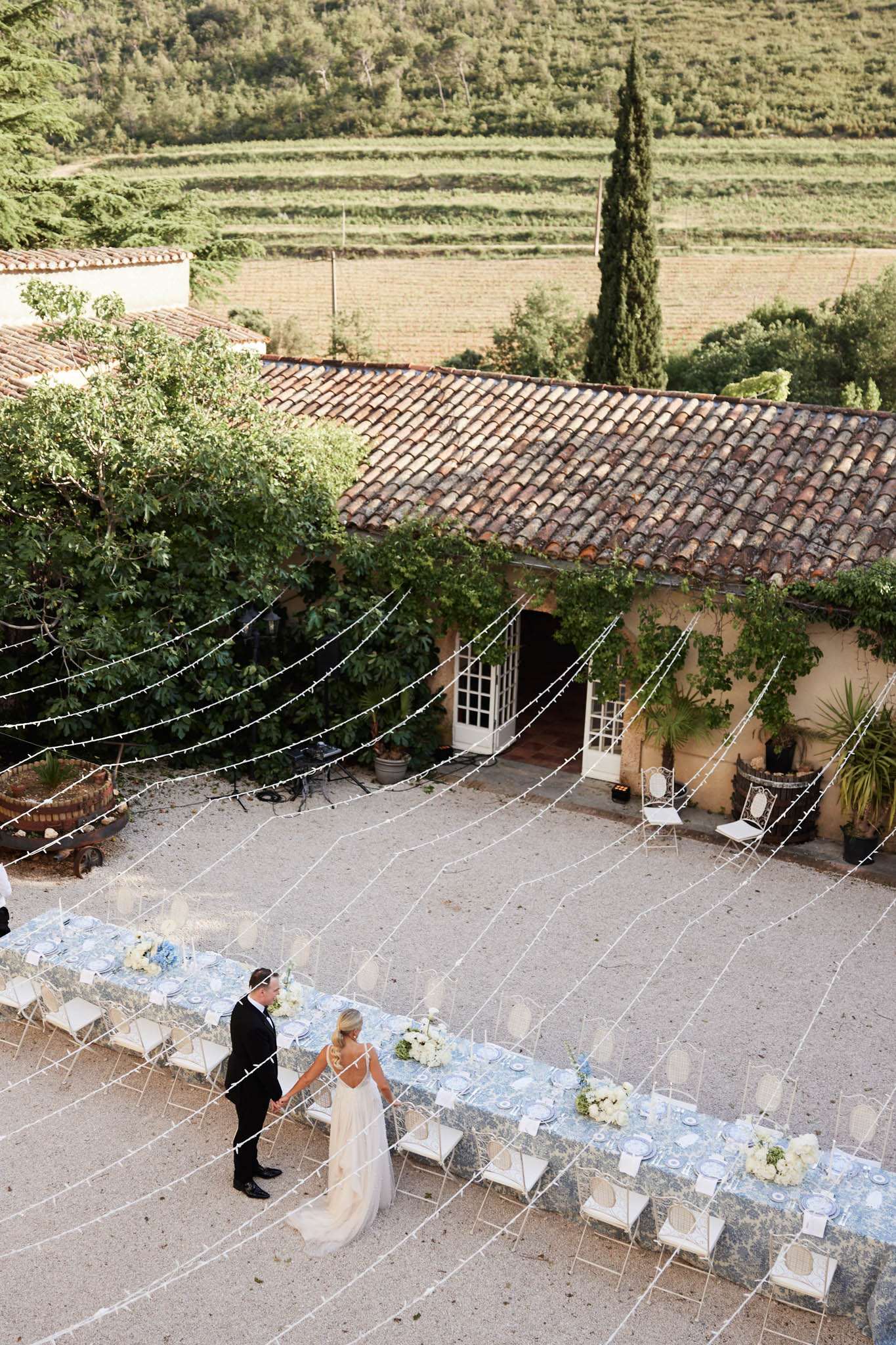 An aerial wide shot captures a bride and groom walking hand in hand through an outdoor courtyard reception setup at a Provençal farmhouse or mas, surrounded by terracotta-tiled rooflines and vineyard terraces in the background. The bride wears a low-back ivory gown with a train, and the groom is dressed in a black tuxedo. Two long banquet tables run parallel across the gravel courtyard, dressed in blue-and-white toile or floral-print linens, set with blue-rimmed plates and centerpieces of white and pale yellow florals including hydrangeas and roses with greenery. White iron chairs line both tables, and white fairy lights are strung in arching lines across the courtyard overhead. The overall styling is classic French country with a blue-and-white palette and intimate scale. Potential venue feature image.