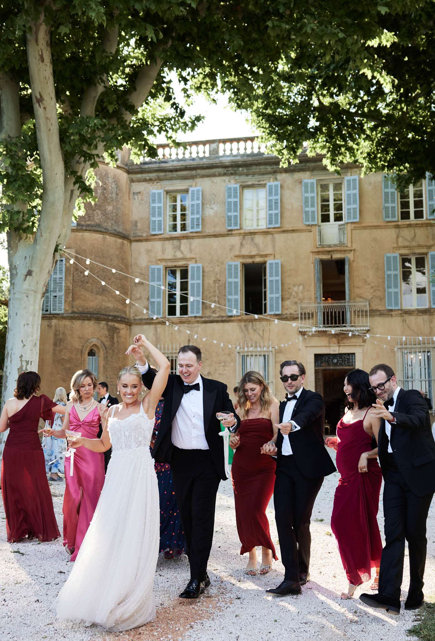 The bride and groom are dancing and celebrating outdoors on a gravel courtyard in front of a classic French château with warm golden stone façade, pale blue shutters, and a wrought iron balcony. The groom, in a black tuxedo with bow tie, holds the bride's hand raised as they dance, while the bride wears a spaghetti-strap A-line gown with a beaded bodice. They are surrounded by approximately six guests who are also dancing and laughing; several women wear deep burgundy and hot pink floor-length gowns, and the men are dressed in black suits and tuxedos. String globe lights are strung across the courtyard between the trees, adding to the festive outdoor reception atmosphere. The image is a candid wide shot capturing a lively, joyful moment with the château façade prominently visible in the background. Potential venue feature image.