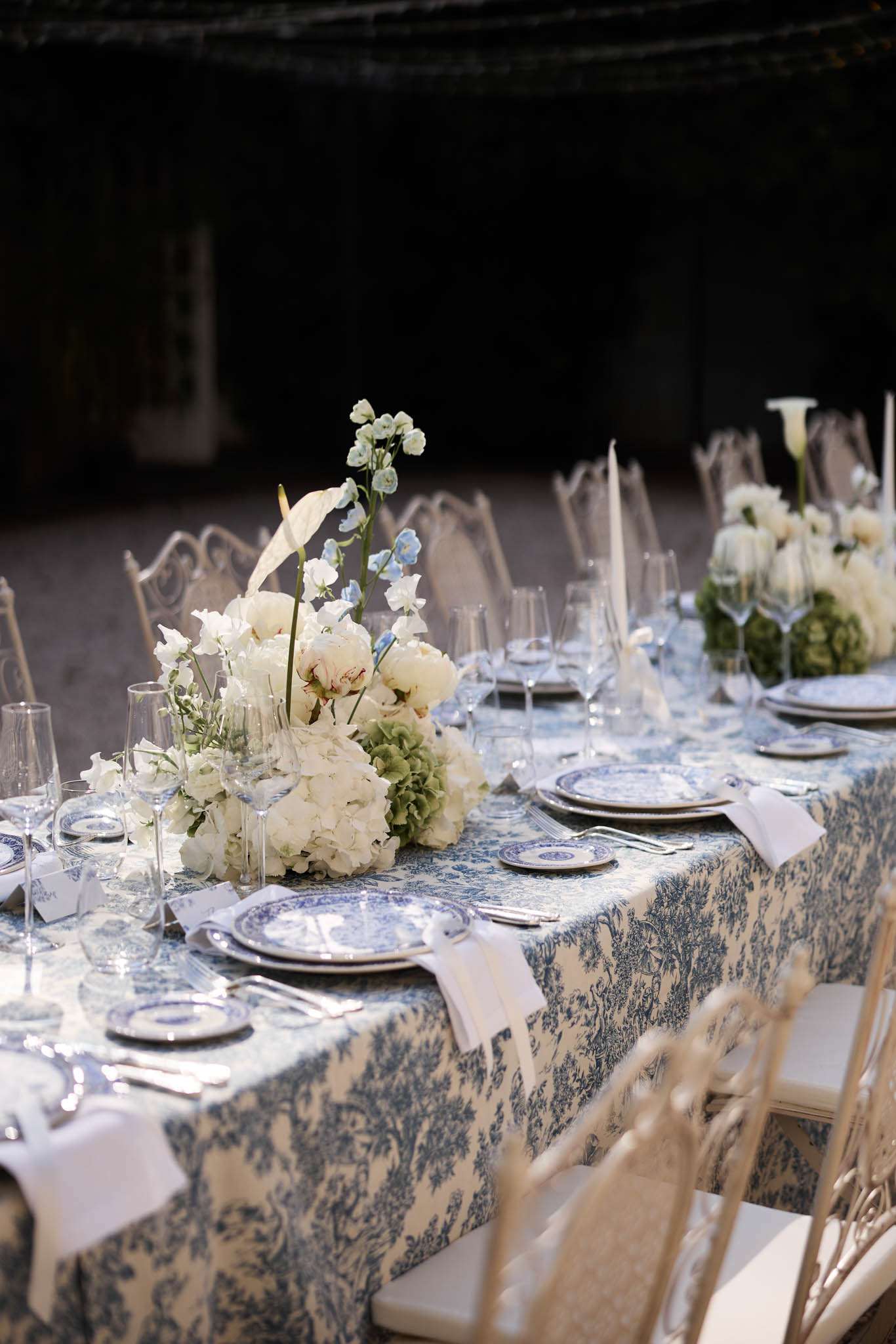 A detail shot of a styled wedding reception table set outdoors, photographed at an angle to show the full length of the setting. The table is covered in a blue-and-white toile de Jouy patterned tablecloth, topped with blue-and-white patterned charger plates stacked with matching dinner plates, white linen napkins, silver cutlery, and multiple clear crystal wine and champagne glasses per place setting. The centerpieces consist of low, lush arrangements featuring ivory peonies, white hydrangeas, green hydrangeas, white sweet peas, pale blue delphiniums, and white anthurium leaves, with a taller calla lily arrangement visible further along the table. Gold chiavari chairs are positioned along both sides of the table. The overall decor palette is blue, white, and soft green, in a classic French-inspired style.
