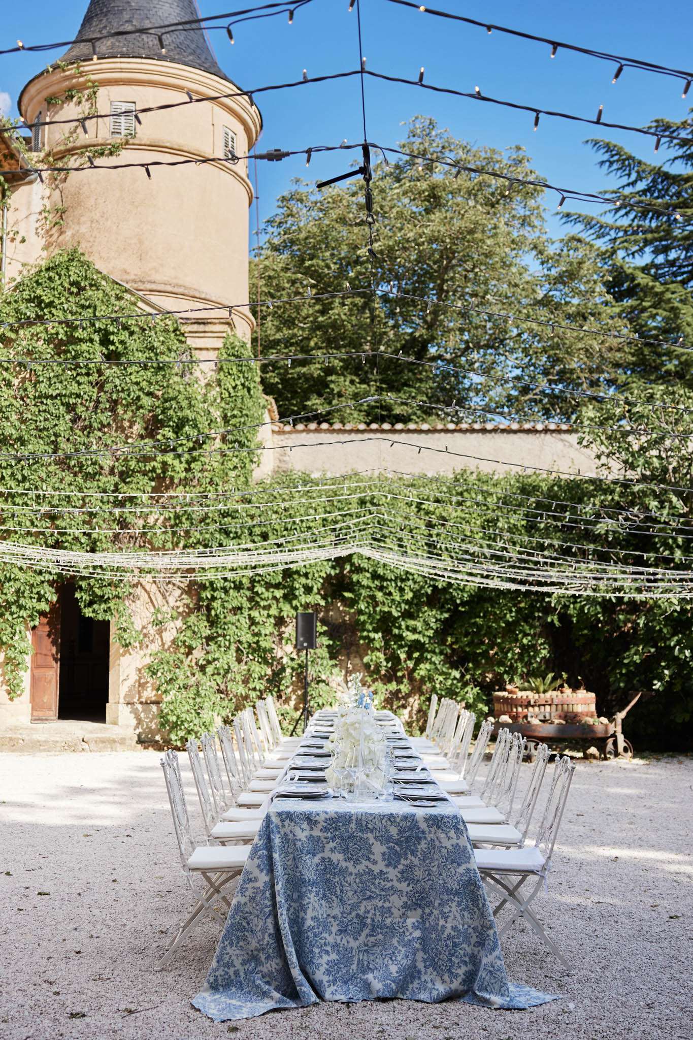 An outdoor wedding reception table is set up in the courtyard of a French château, featuring a long rectangular table draped in a blue-and-white damask-patterned tablecloth. The table is lined on both sides with white folding chairs with padded seats, and centrepieces of white flowers — appearing to be hydrangeas or similar blooms — are placed along the centre alongside glassware and dark charger plates. Overhead, strings of bistro-style globe lights and what appear to be hanging white floral garlands are suspended across the courtyard on black cables, creating a canopy effect. The setting is a wide establishing shot showing the full table length receding toward the château's ivy-covered stone tower with a dark conical roof in the background. Potential venue feature image.