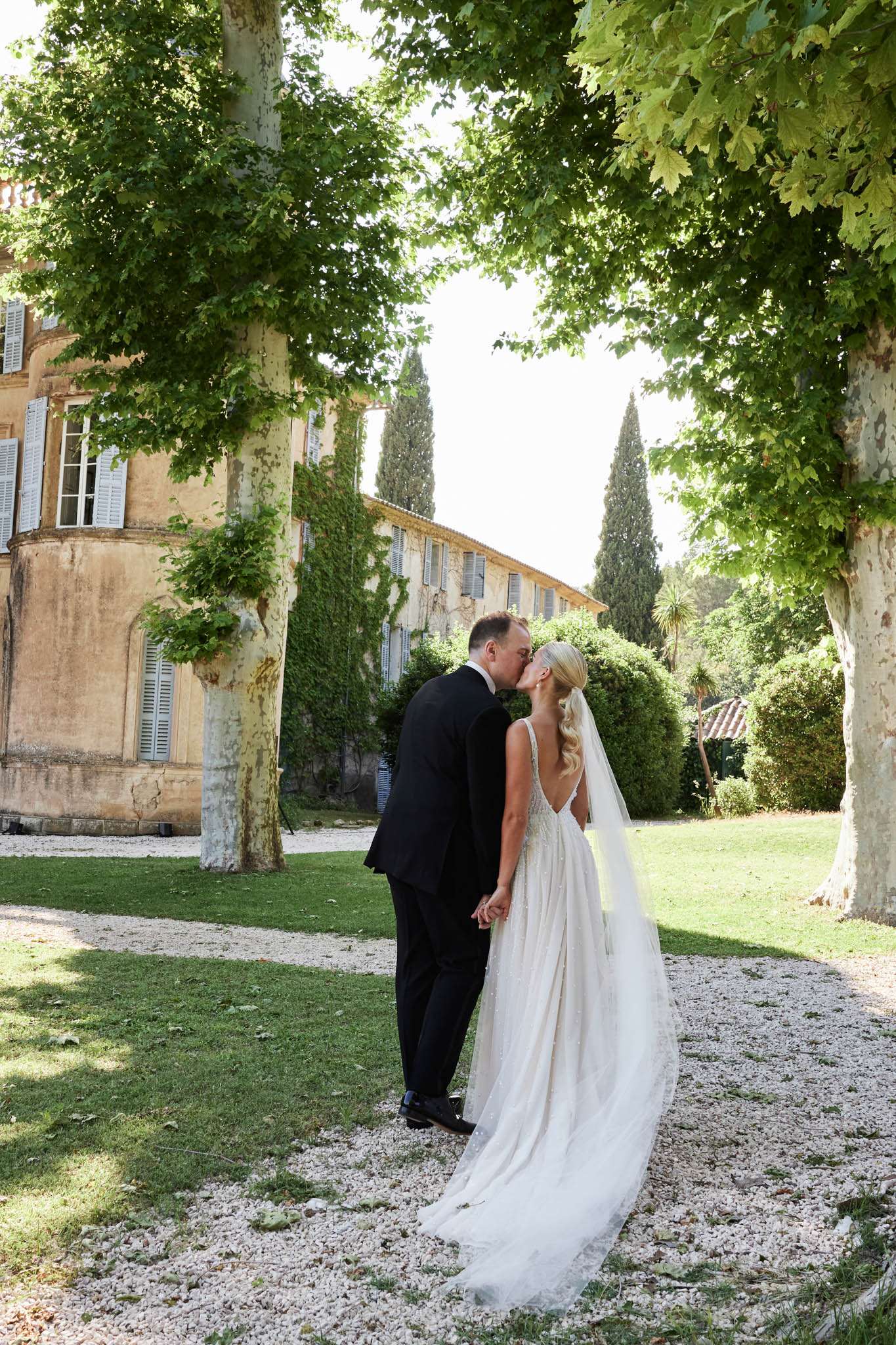 Couple kissing on chateau gravel grounds bride in low-back gown with cathedral veil ochre Provencal facade behind