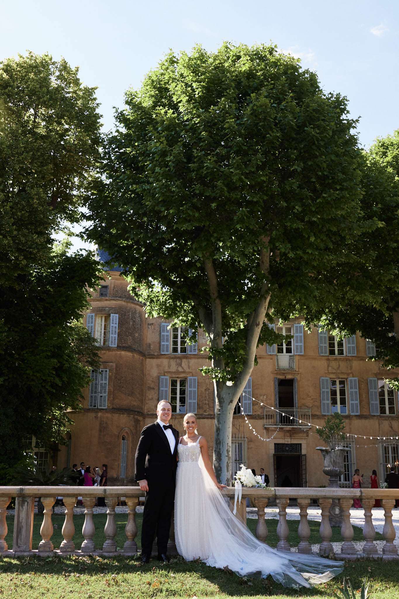 Bride in lace gown with cathedral train and groom in black tuxedo posing before honey-stone chateau