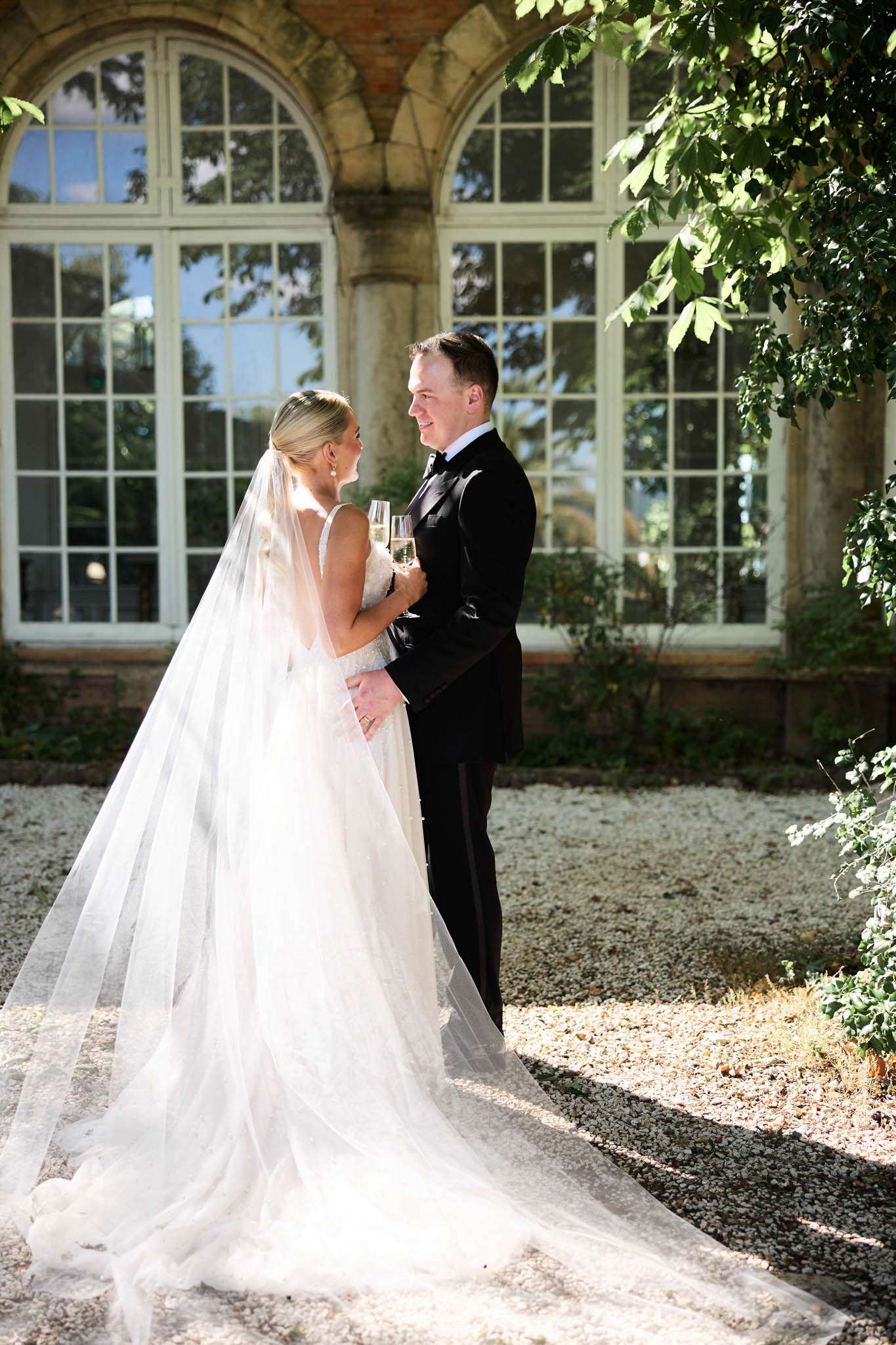 Bride in V-neck ballgown with cathedral veil and groom with champagne before stone orangerie arch