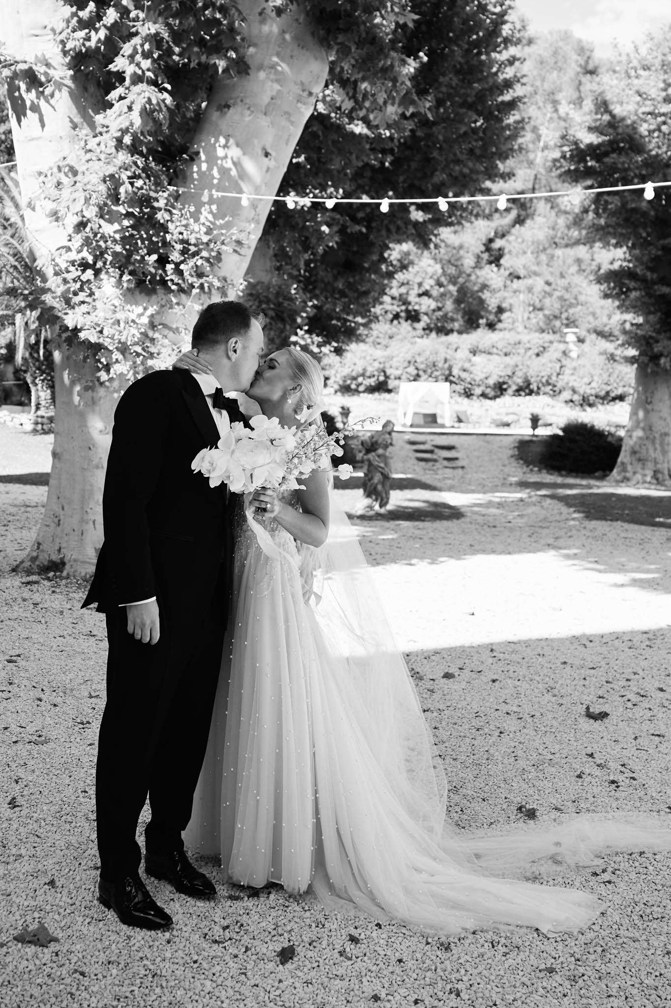 Black and white couple kissing on gravel with beaded ballgown train and globe lights in trees